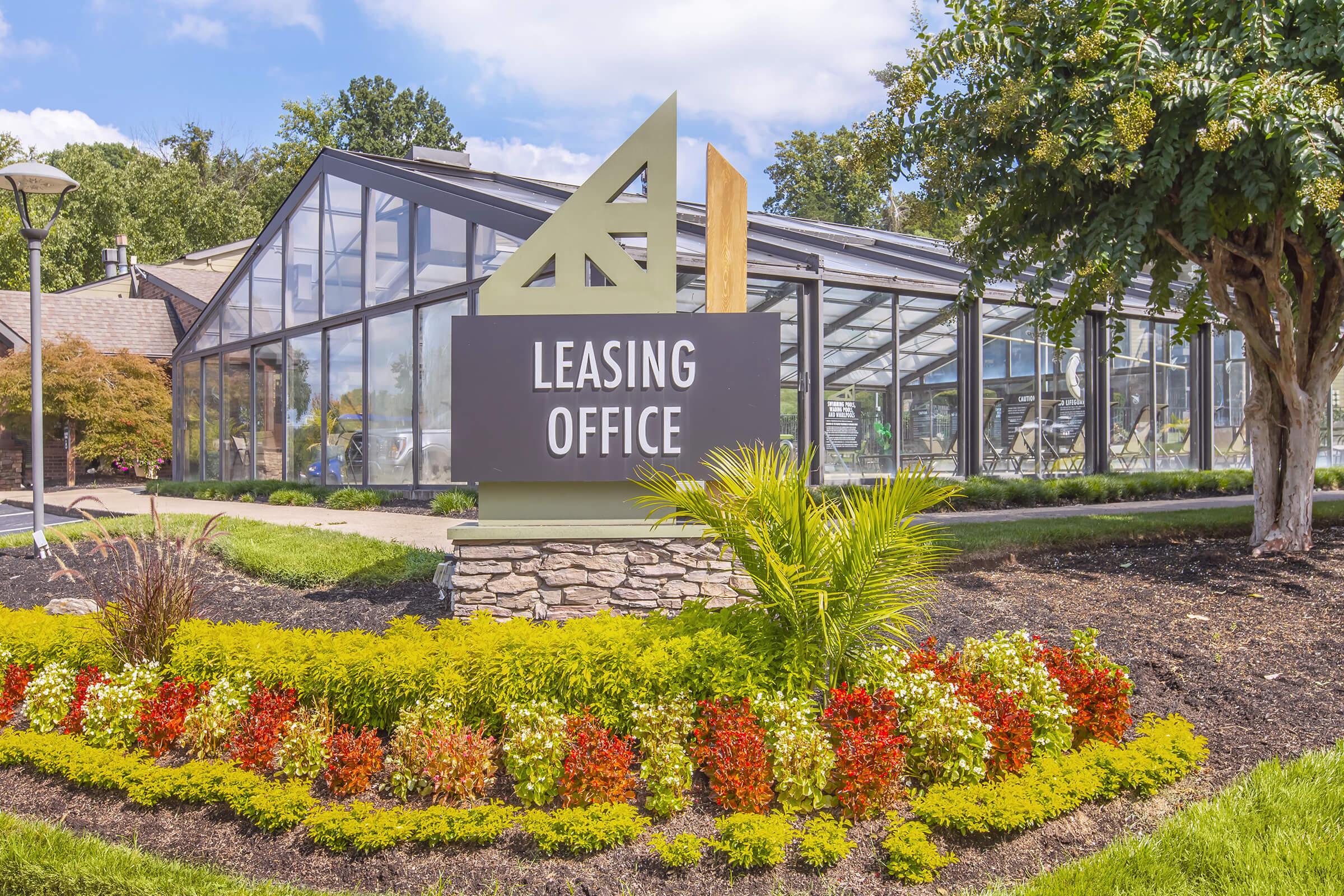 Leasing office building surrounded by well-maintained gardens featuring colorful flower beds and greenery. The structure has large glass windows and a modern architectural design, highlighted by a prominent sign reading "Leasing Office." Clear blue sky and trees are visible in the background.