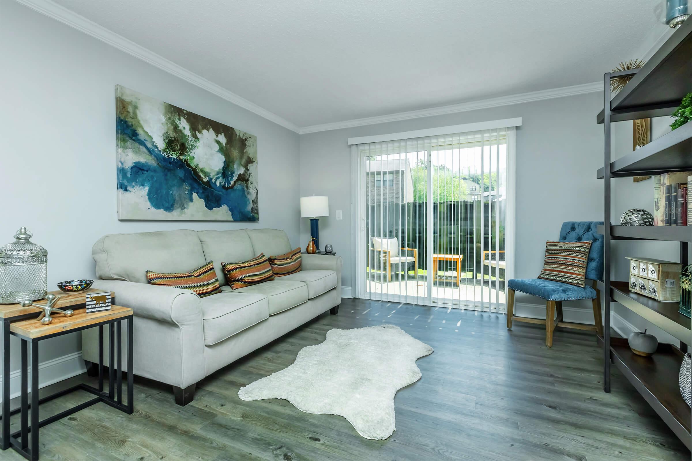A modern living room featuring a neutral-colored couch with decorative cushions, a wooden side table with a decorative bowl, and a light-colored area rug. A large abstract painting in blue and green hangs on the wall. There's a sliding glass door leading to an outdoor area with two chairs visible outside.