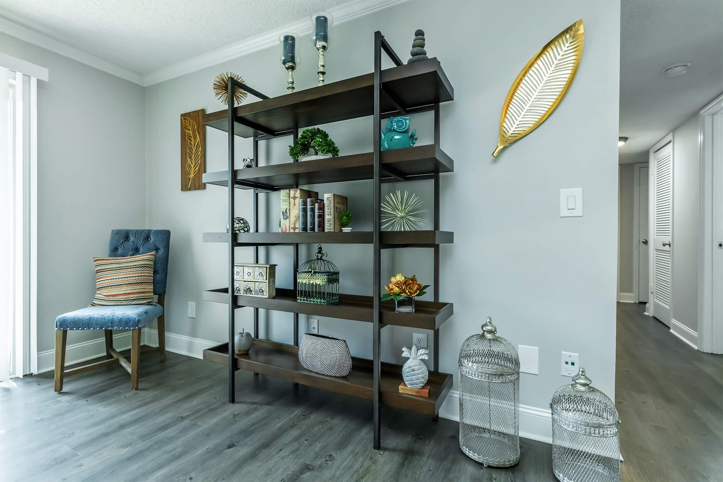 A modern shelving unit with various decorative items, including books, a glass birdcage, and a green vase. The backdrop features a blue chair with striped cushions and decorative wall elements, such as a gold leaf and a geometric design. The room has a neutral color palette and natural light.