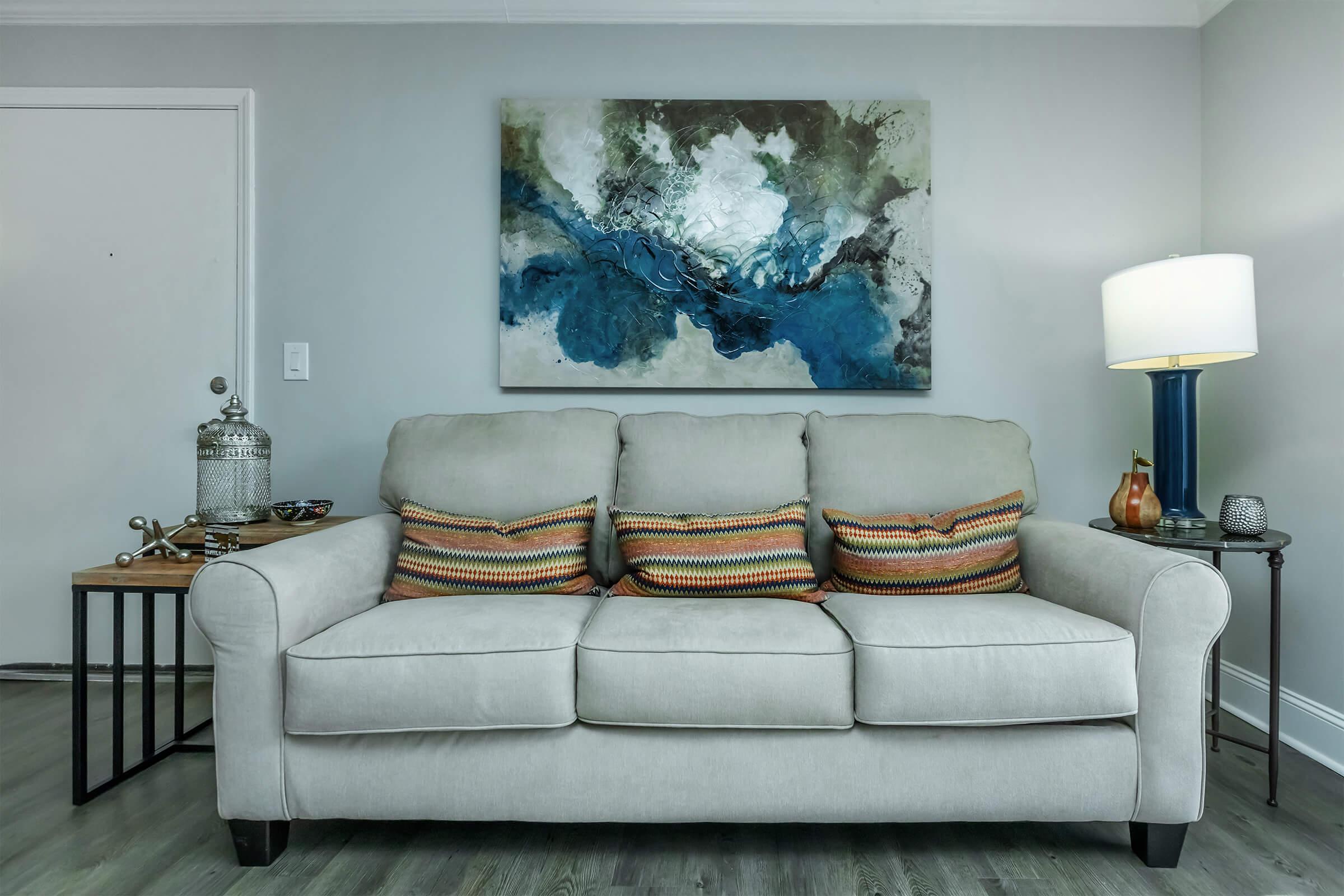 A light beige sofa with colorful striped cushions is positioned in a modern living room. Next to it, there's a small wooden side table with various decorative items. A large abstract painting featuring blue, green, and white hues hangs on the wall above the sofa, while a lamp stands on the opposite side, providing warm light.