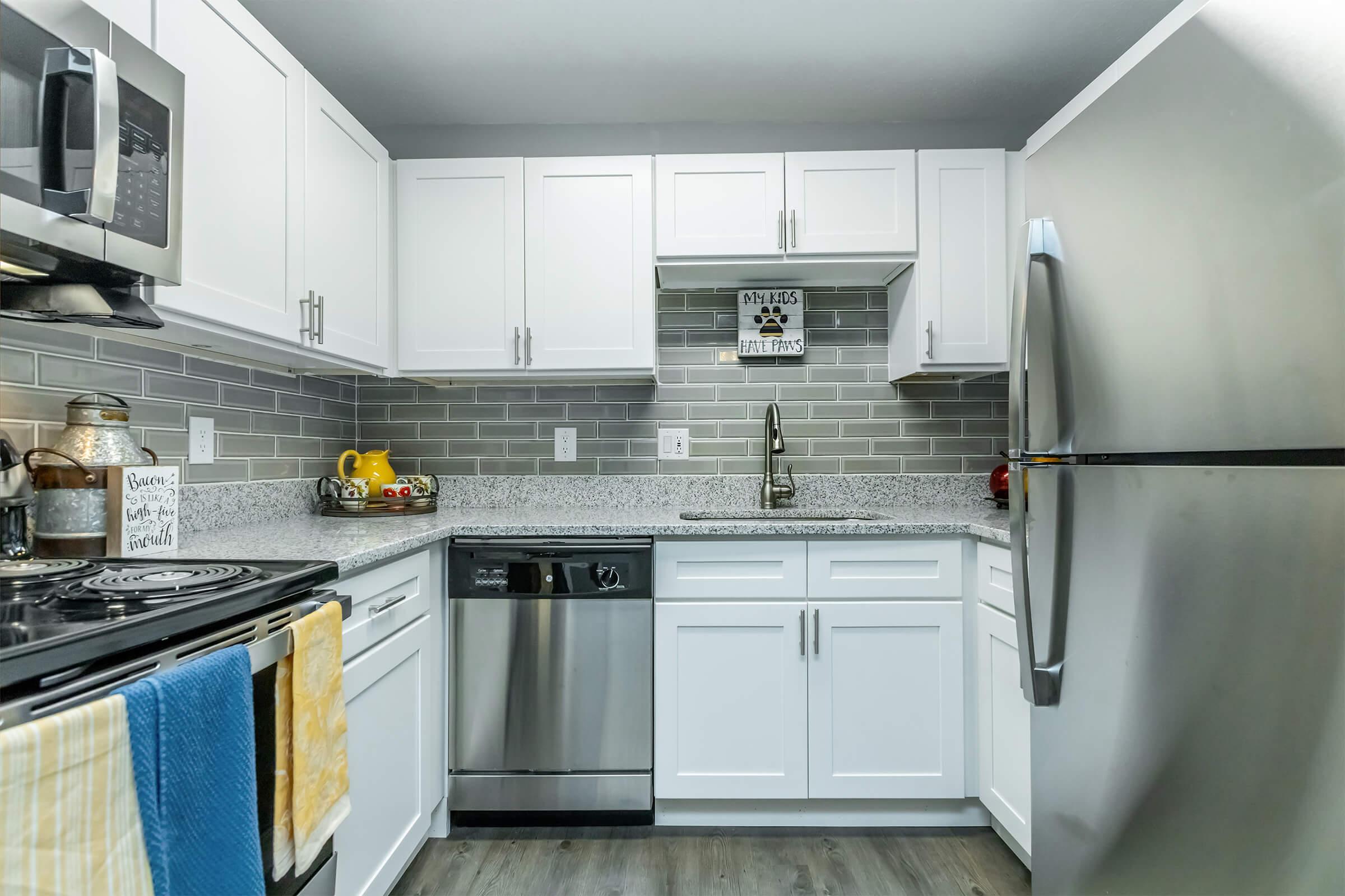 Modern kitchen featuring white cabinetry and gray backsplash tiles. Stainless steel appliances including a refrigerator and dishwasher. The countertop is gray speckled material, and there are decorative items like fruits and a sign on the wall. Soft lighting adds a warm ambiance.