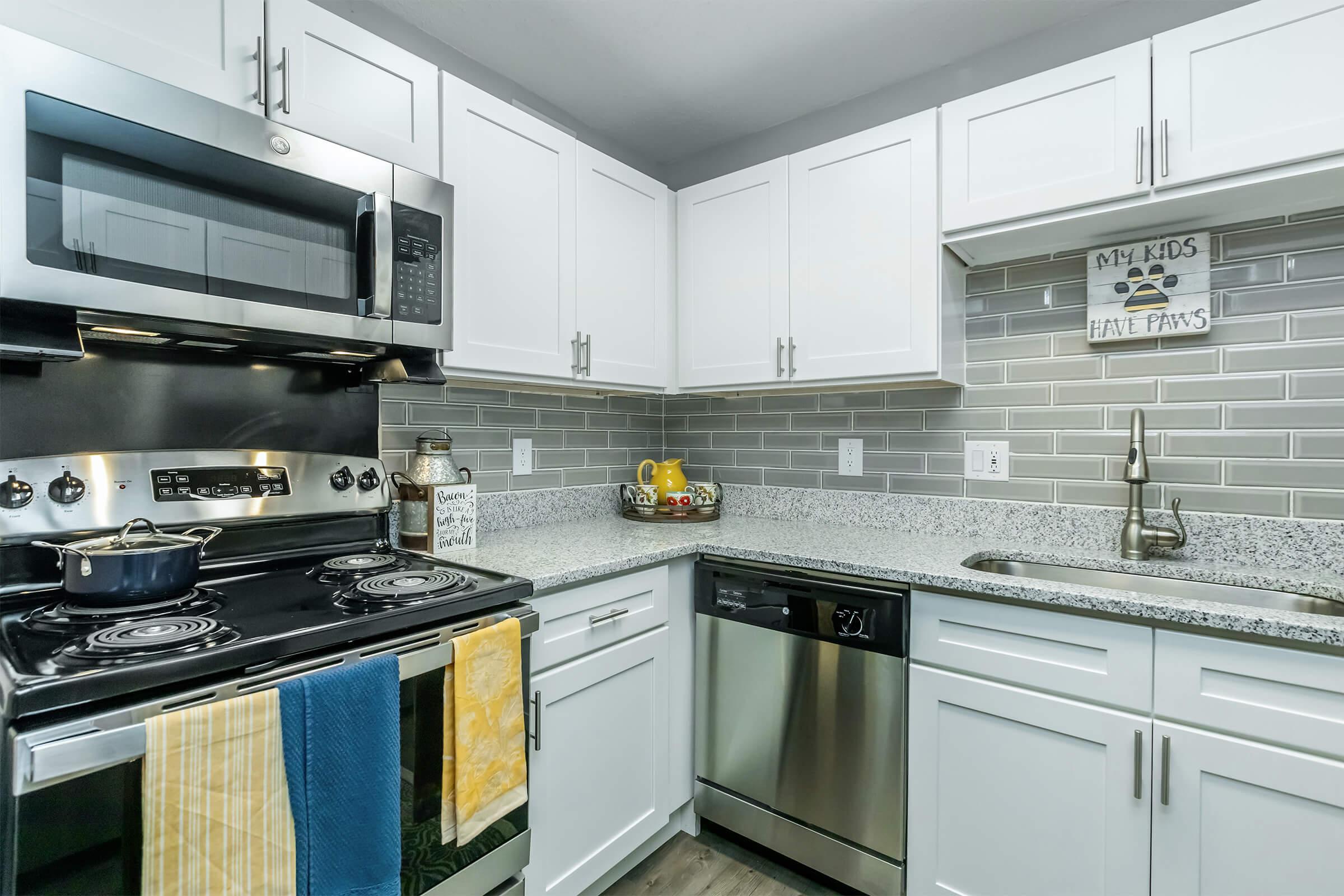 Modern kitchen featuring stainless steel appliances, including an oven, microwave, and dishwasher. The countertops are gray speckled granite with under-cabinet lighting. Gray tiled backsplash with decorative signs and a bowl of fruit on the counter. Towels hang from the stove, adding a pop of color.