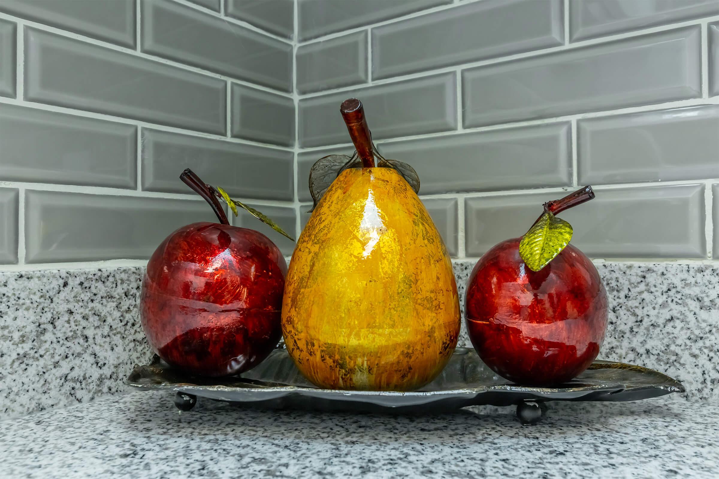 A decorative display featuring three artificial fruits: two red apples and one yellow pear, arranged on a silver tray. The backdrop consists of gray tiled walls, and the fruits are placed on a granite countertop.