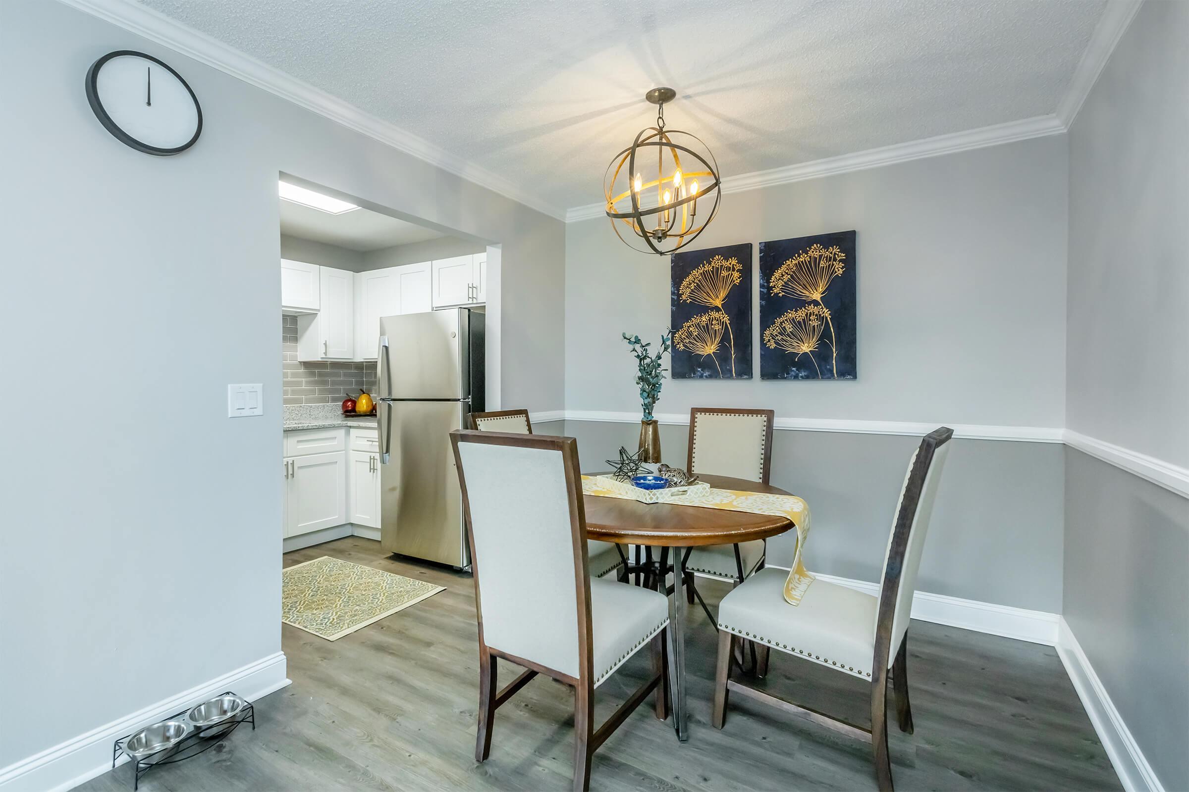 A modern dining area featuring a round table with four upholstered chairs. On the table sits a decorative plate and a small vase with greenery. The walls are painted in light gray, complemented by a wainscoting detail. A stylish light fixture hangs above, and a clock is mounted on the wall. A kitchen is visible in the background.