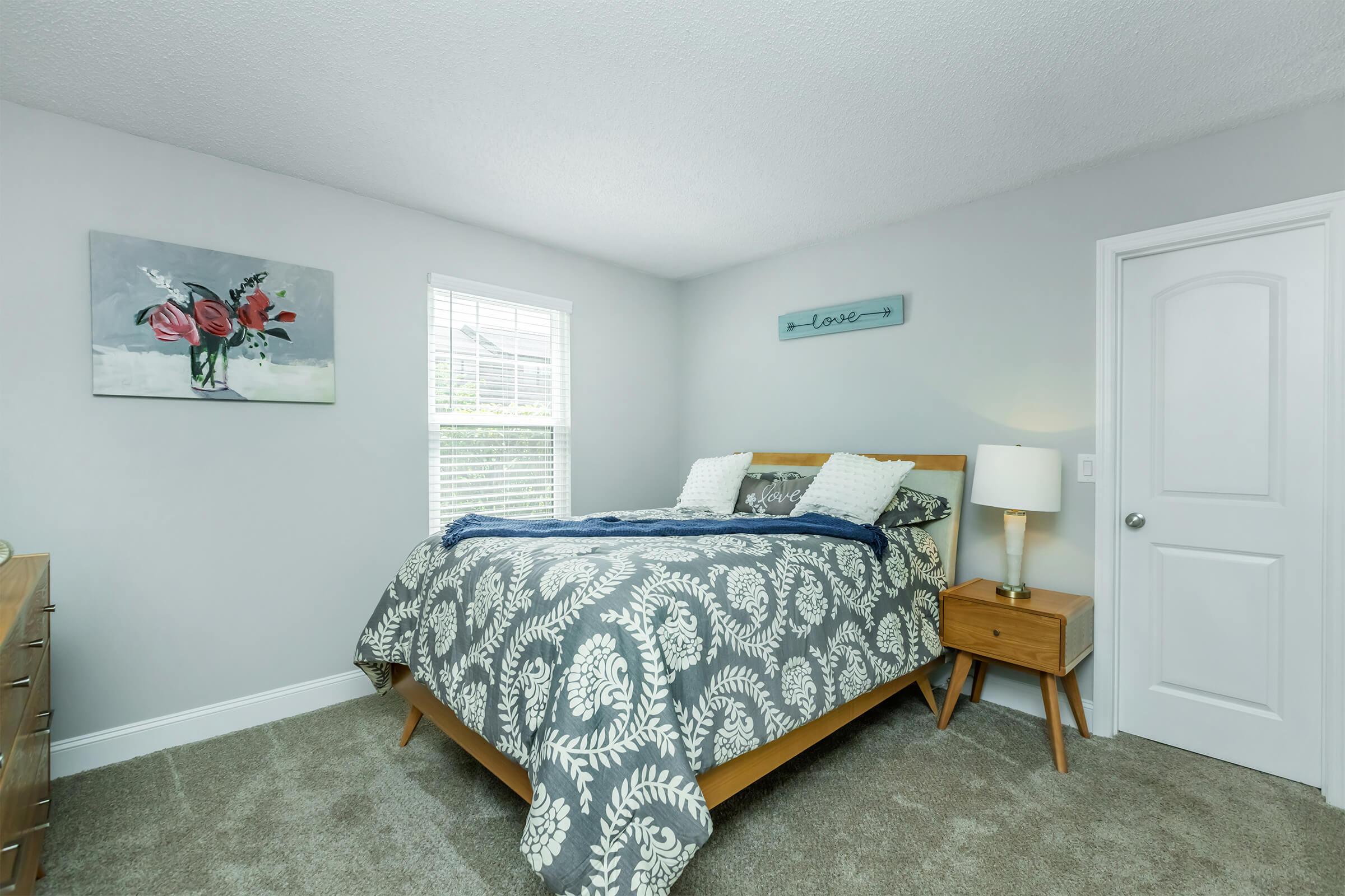 A cozy bedroom featuring a wooden bed with a patterned gray and white blanket. A nightstand with a lamp sits beside the bed. There is a painting of flowers on the wall and a small sign that says "love." Bright natural light filters through a window, enhancing the warm atmosphere.
