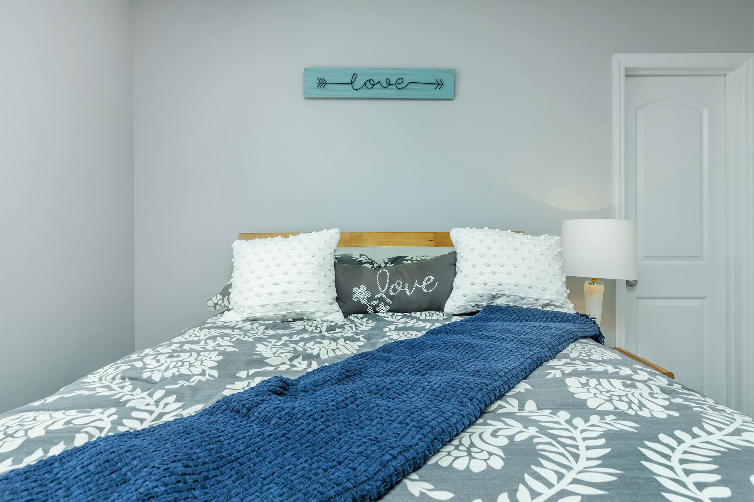 A cozy bedroom featuring a bed with a patterned gray comforter and white pillows. Two decorative pillows with the word "love" are on the bed. A blue knit throw blanket is draped across the foot of the bed. A bedside lamp is visible along with a light gray wall and a decorative sign above the bed that says "love."