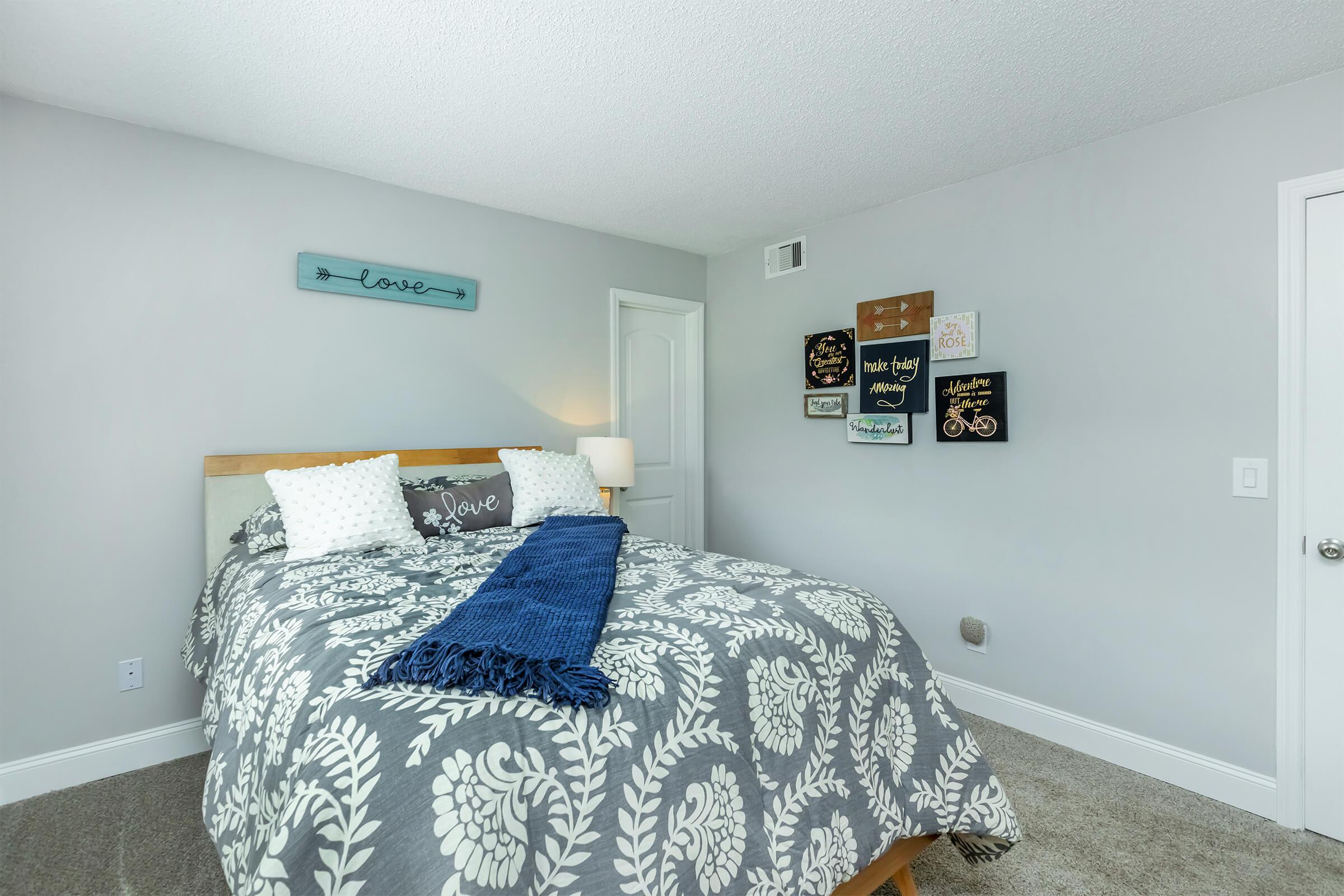 A cozy bedroom with a light gray color scheme, featuring a neatly made bed with a patterned gray and white comforter. Two pillows with decorative text sit on the bed, and a blue throw blanket drapes over the edge. The walls are adorned with various framed signs and a small "love" decoration above the bed.