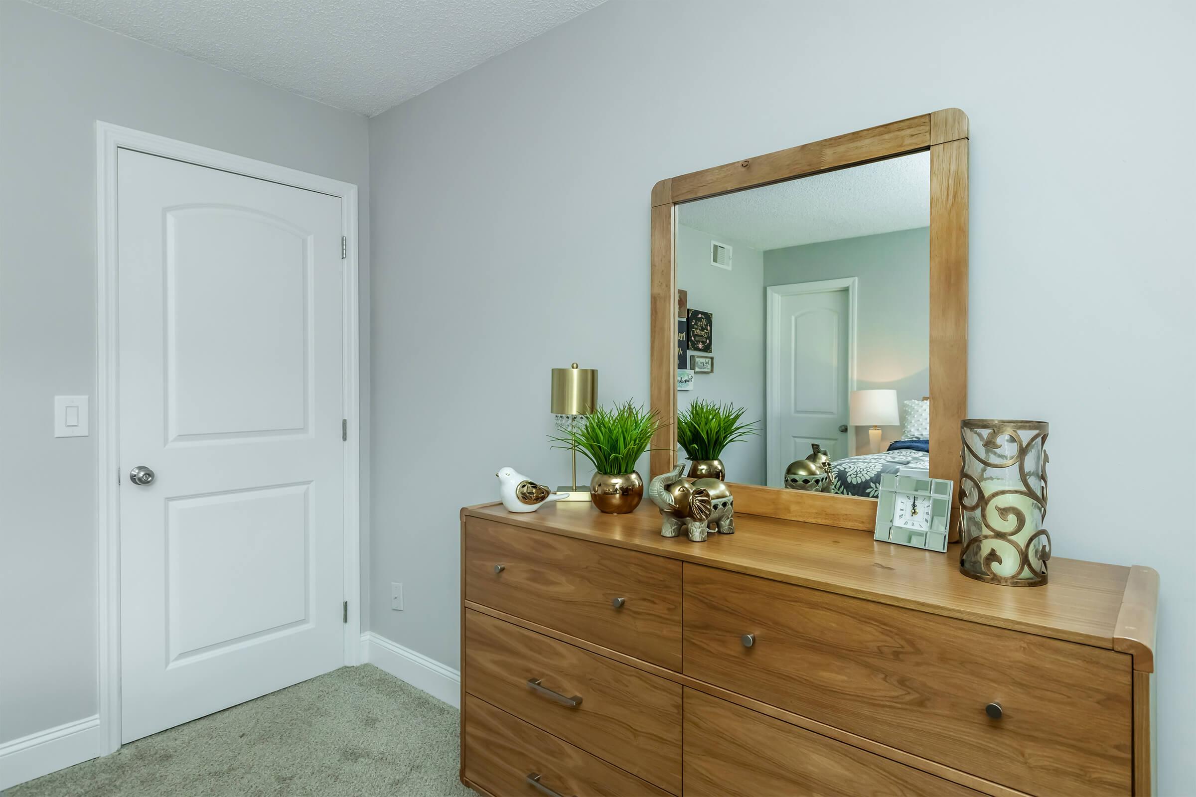 A wooden dresser with a mirror is positioned against a light gray wall in a well-lit bedroom. The dresser features decorative items, including small plants and a picture frame. A white door is visible in the background, along with a glimpse of a bed. The room has a clean and modern aesthetic.