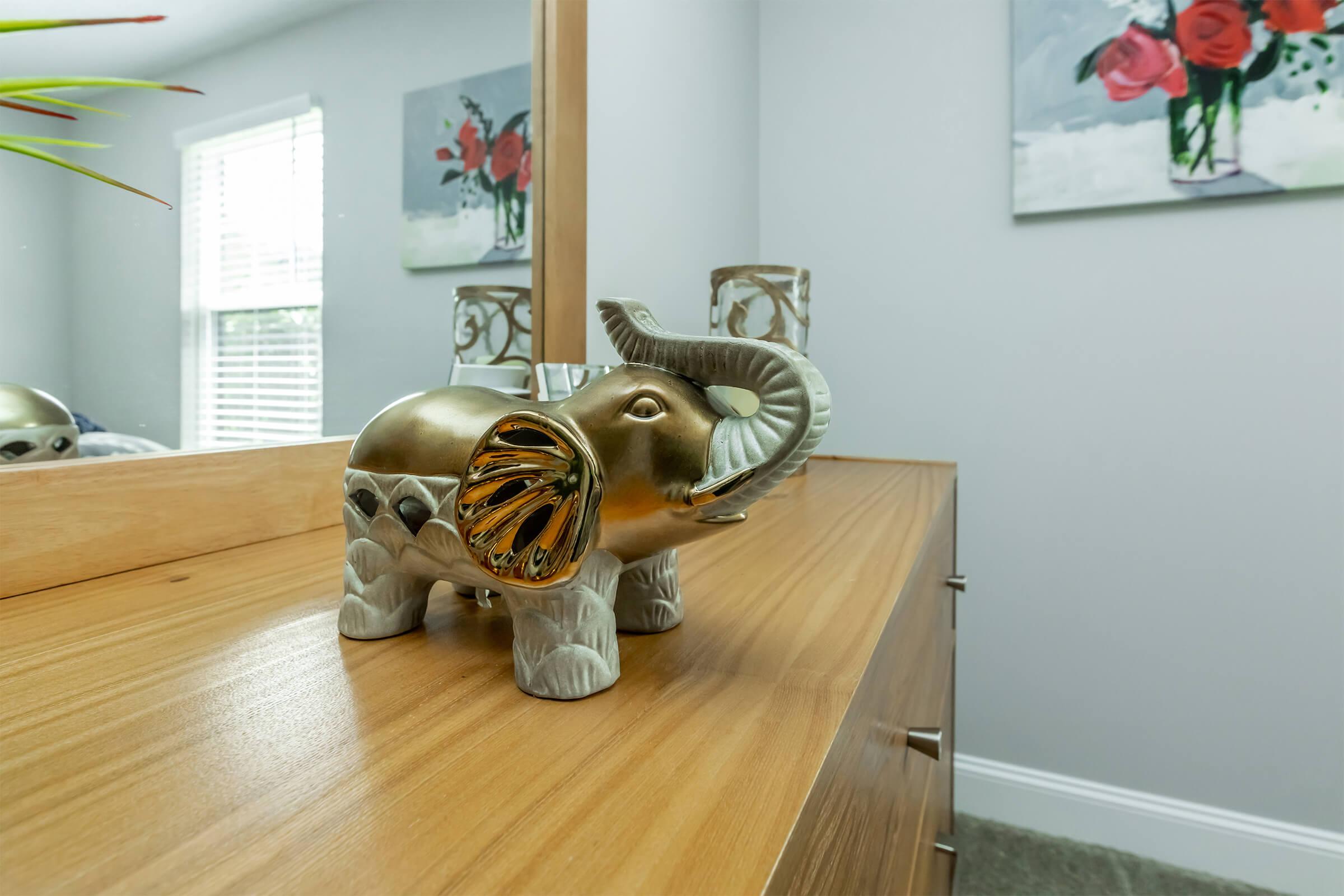 A decorative golden elephant figurine with intricate designs stands on a wooden dresser. In the background, there are two floral paintings and a light-filled room with a window, enhancing the serene ambiance of the space.