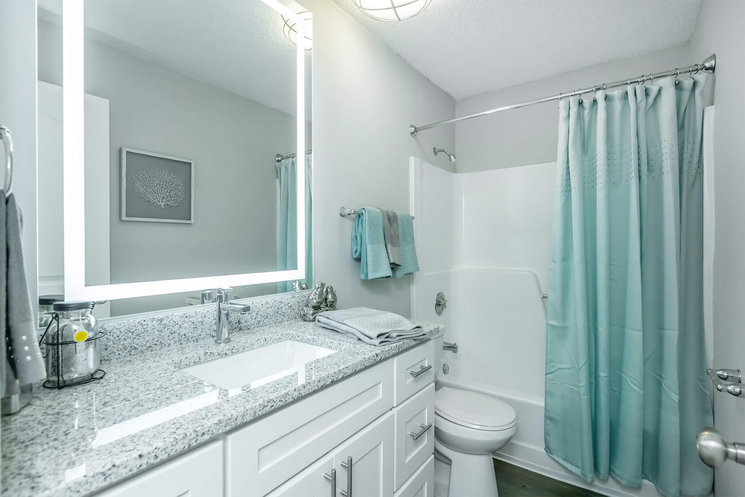 A modern bathroom featuring a freestanding bathtub, a white vanity with a sink and granite countertop, a large illuminated mirror, light grey walls, and a shower with a light blue curtain. Soft towels hang on the vanity, and a small decorative piece is mounted on the wall.