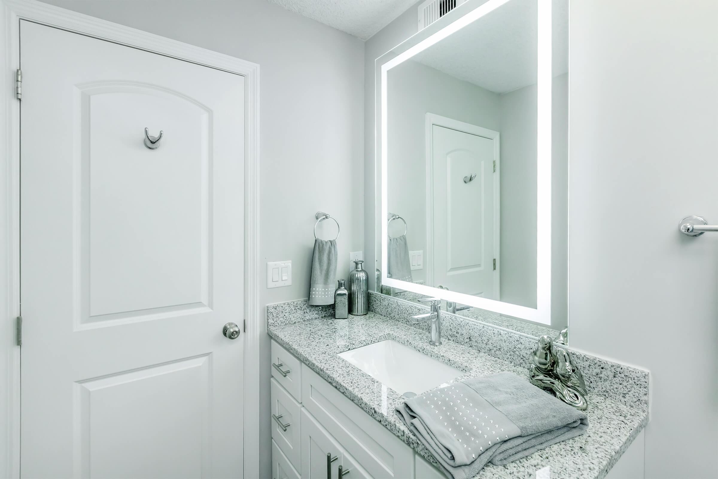 A modern bathroom featuring a sleek white countertop with a sink, illuminated by a large rectangular mirror with built-in LED lighting. The decor is minimalist, with gray towels hanging and light gray walls. A closed door is visible, adding to the clean, contemporary aesthetic of the space.