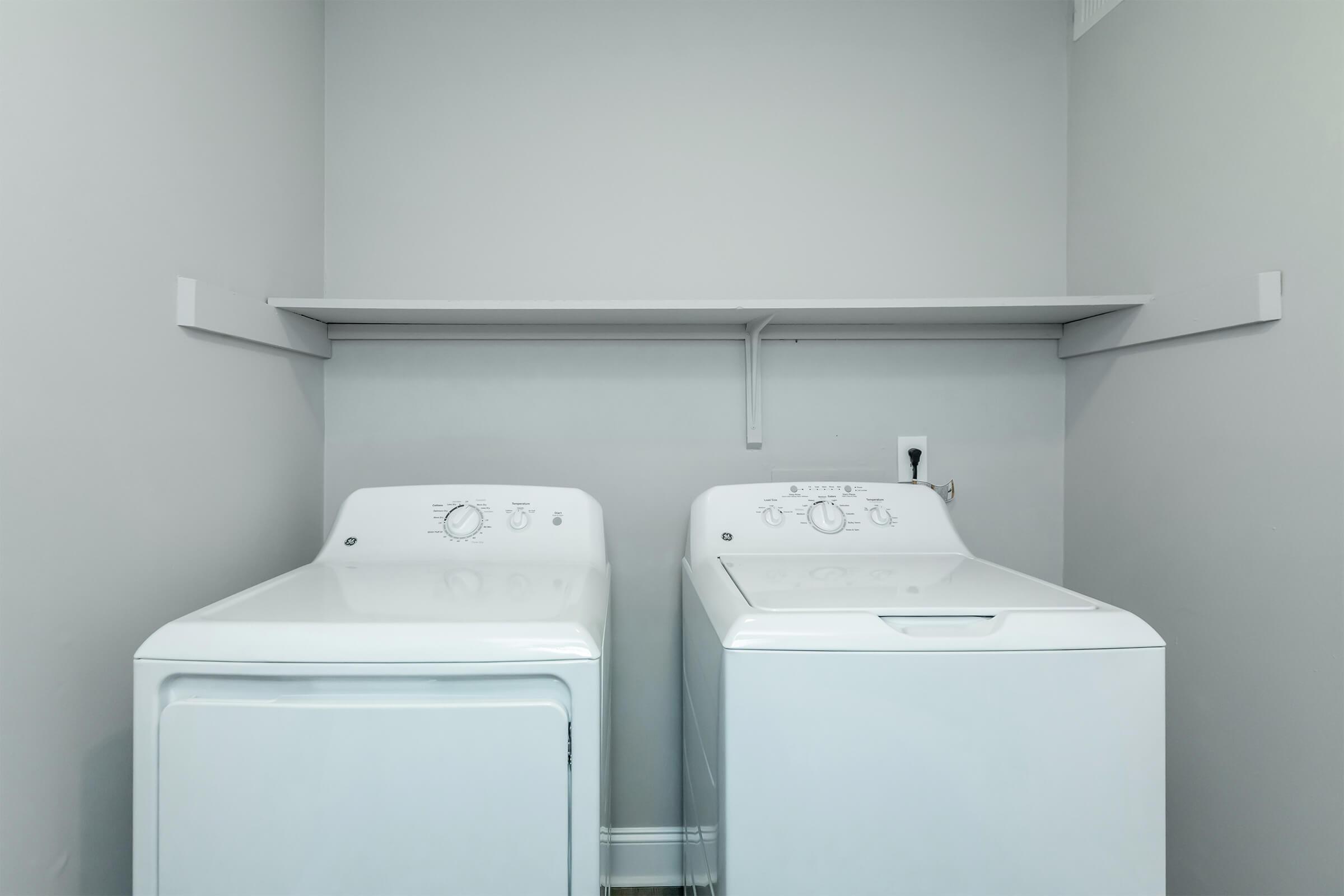 A laundry room featuring a stacked washing machine and dryer in white, placed side by side against a gray wall. Above them, there is a simple shelf for storage. The floor is light-colored, enhancing the spacious feel of the room.