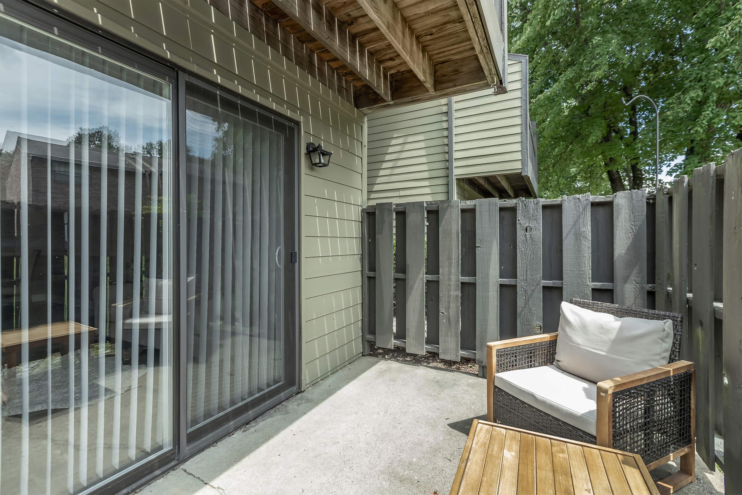 A small outdoor patio area featuring a sliding glass door, a comfortable armchair with a cushion, and a wooden table. The space is enclosed by a dark wooden fence and surrounded by greenery, creating a cozy atmosphere. Note the slanted roof overhead and the natural light filtering in.