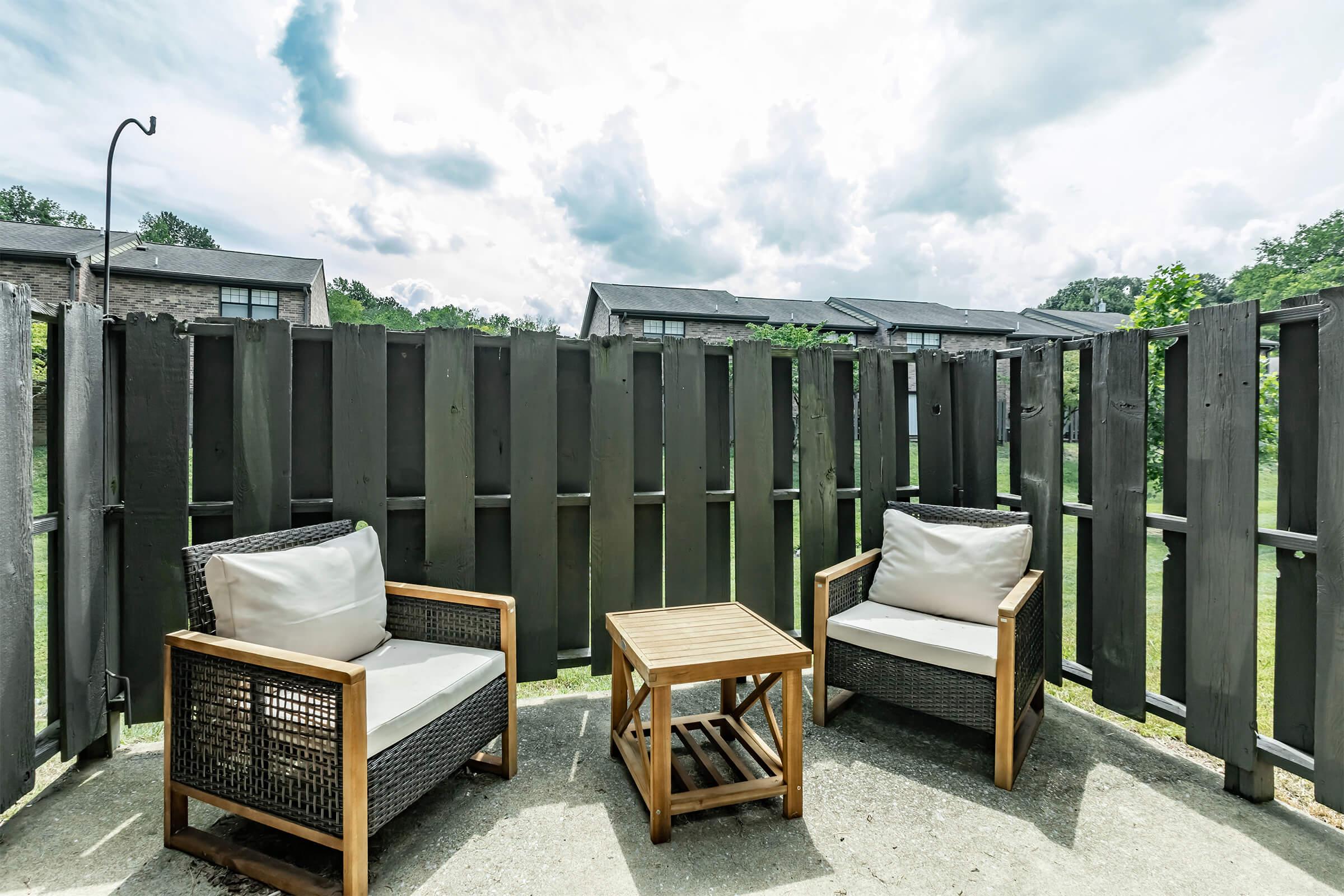 A cozy outdoor seating area featuring two wicker chairs with cushions and a wooden coffee table. The space is enclosed by a wooden fence and is surrounded by green trees and a cloudy sky, creating a peaceful atmosphere.