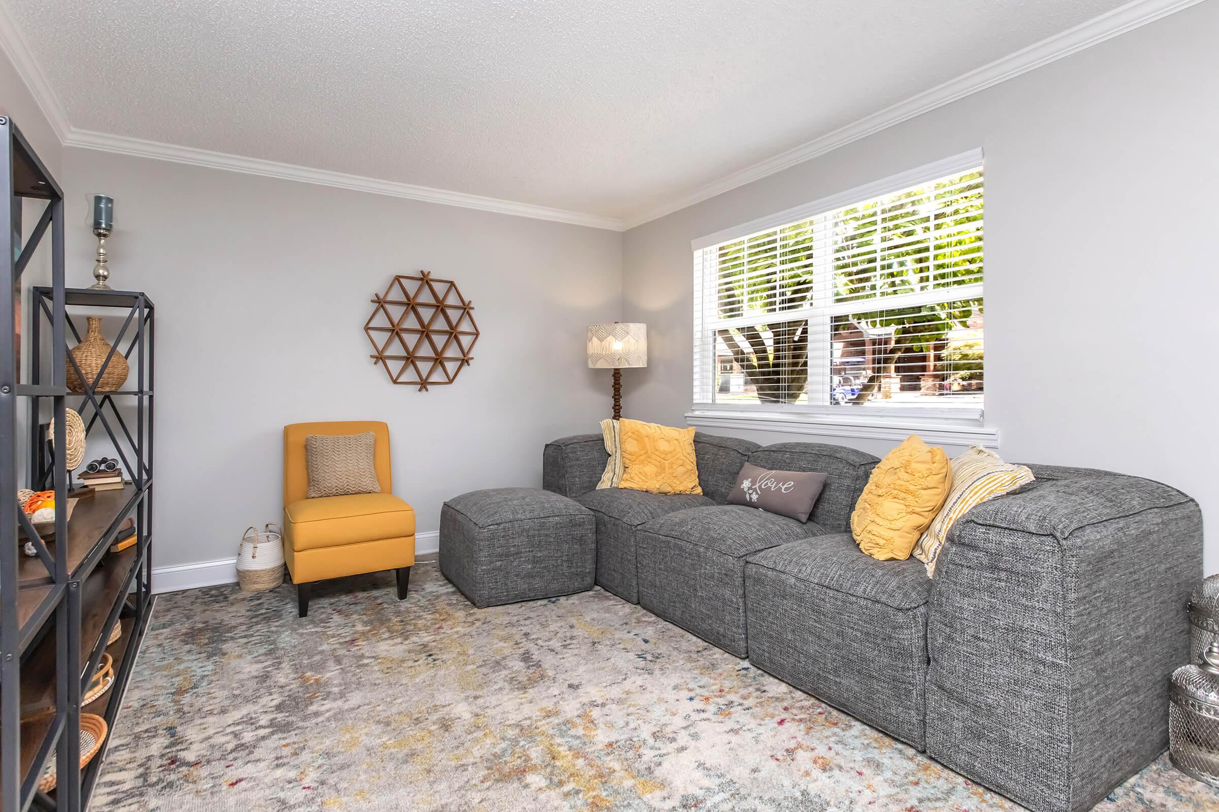 A cozy living room featuring a gray sectional sofa adorned with decorative pillows, a yellow accent chair, a textured area rug, and a stylish wall decor piece. Natural light streams in through the windows, enhancing the inviting atmosphere. A black shelving unit displays decorative items and plants.