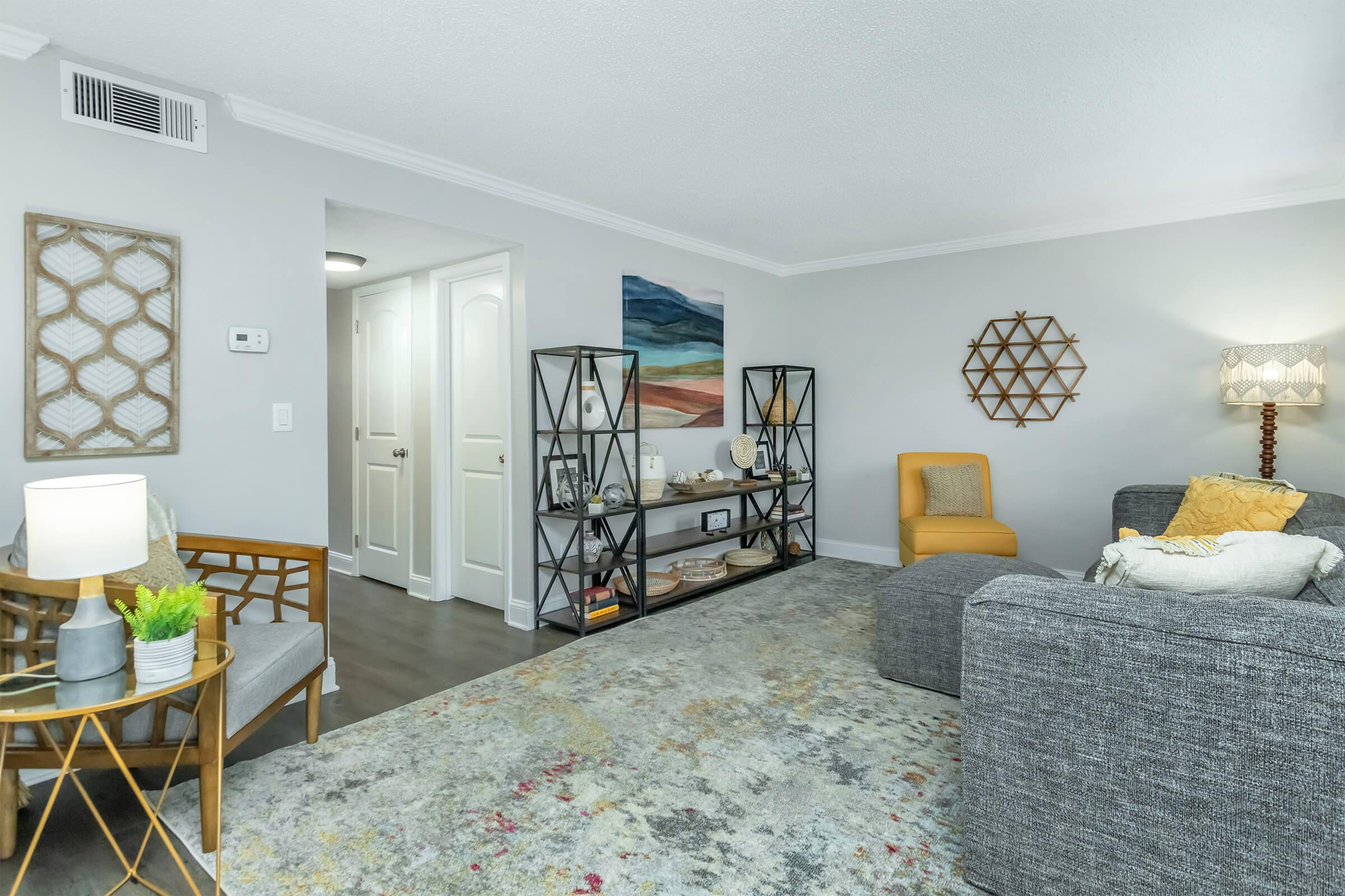 A modern living room featuring a gray sectional sofa, a yellow accent chair, and a decorative rug. There are geometric wall decorations, a lamp, and a stylish bookshelf with decorative items. A doorway leads to another room, and there are colorful artworks on the walls, creating a cozy and inviting atmosphere.