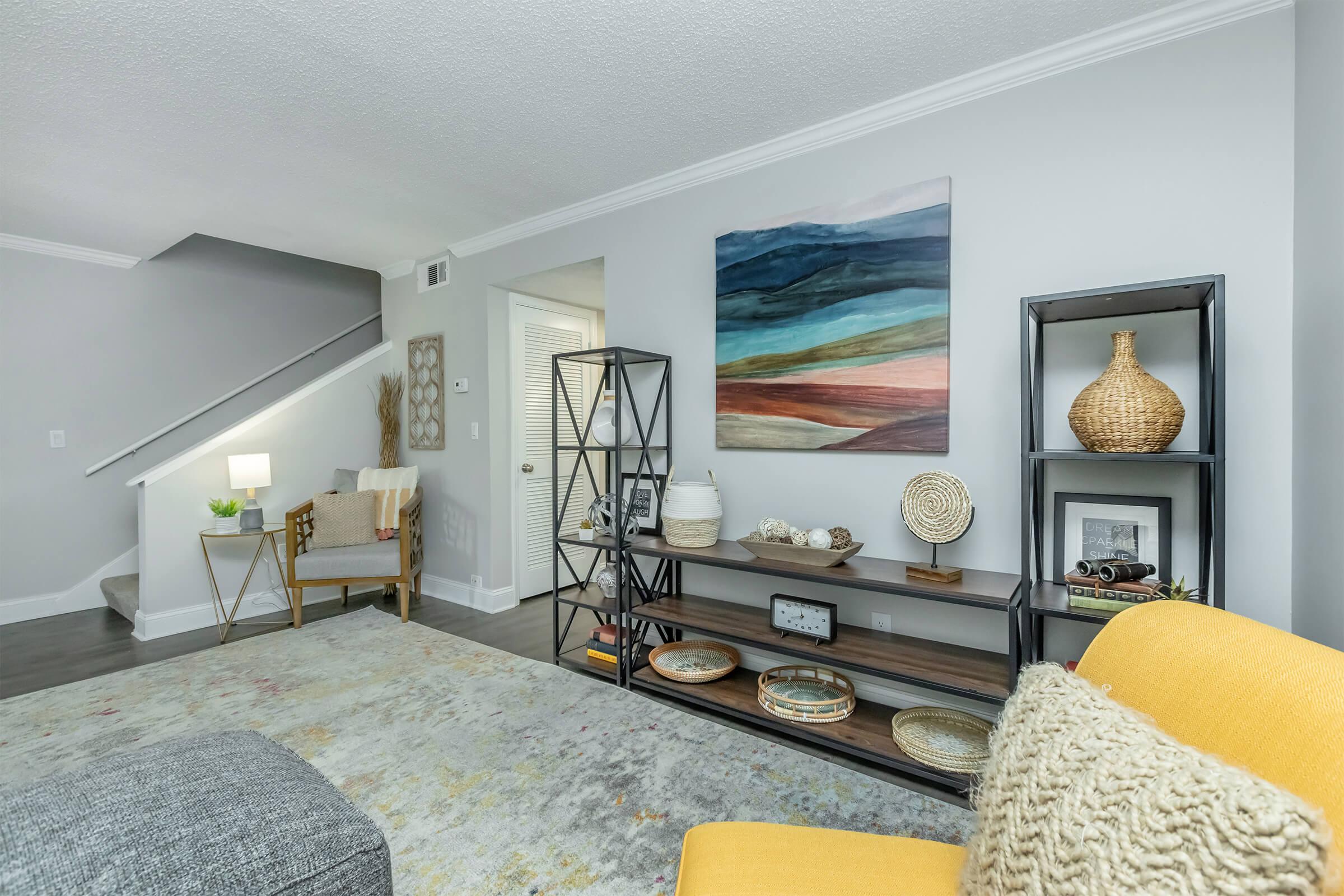 A cozy living room featuring a yellow accent chair, a modern bookshelf with decorative items, and a colorful abstract painting on the wall. A soft area rug covers the floor, and a light fixture illuminates the space. A staircase visible in the background adds depth to the scene.