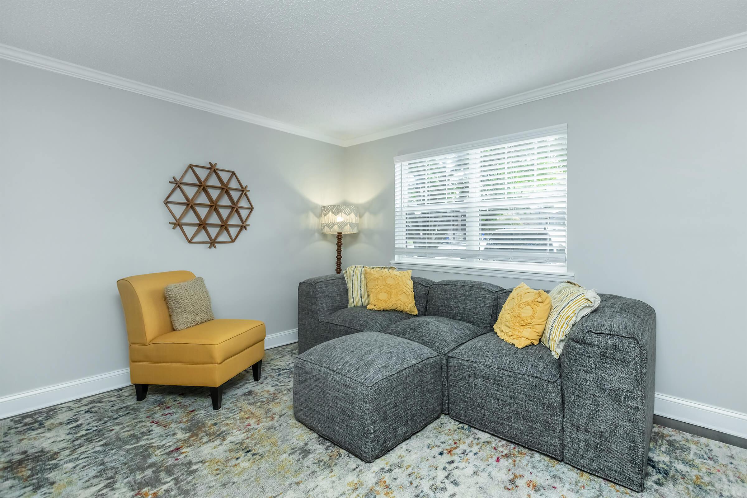 A cozy living room featuring a grey sectional sofa with yellow accent pillows, a yellow accent chair, a geometric wall decor piece, a standing lamp, and a colorful area rug, all set against light grey walls and a window with blinds.
