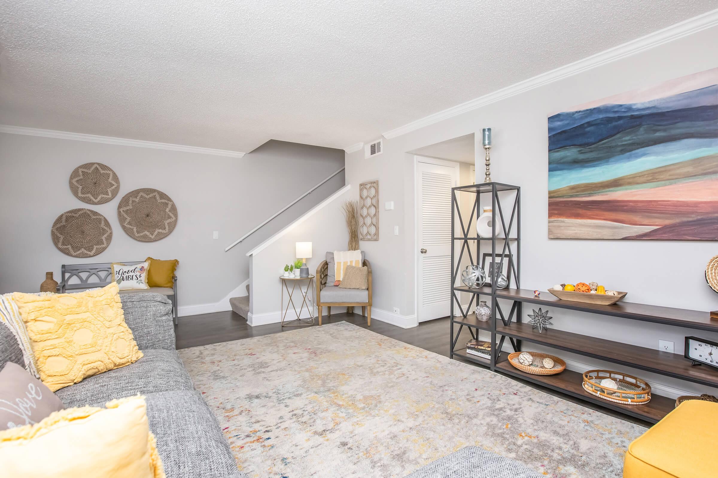 A cozy living room with a gray sofa adorned with yellow pillows. A decorative rug covers the floor, and a modern bookshelf displays various decor items. A colorful painting hangs on the wall, and there are light fixtures and potted plants, creating a welcoming atmosphere. A staircase leads to an upper level.