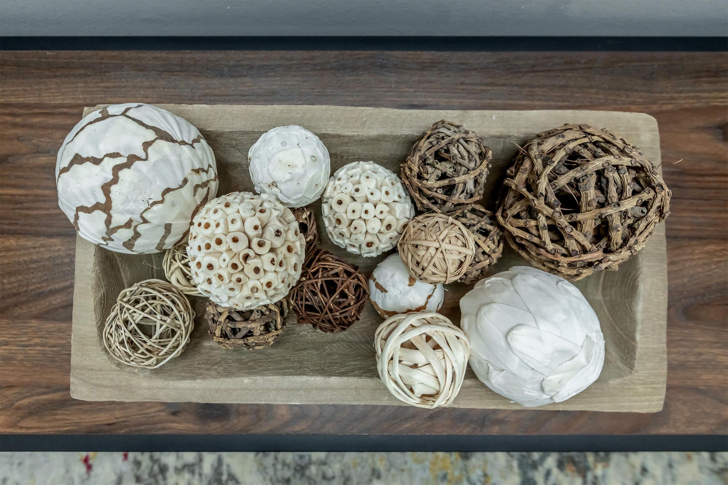 A decorative arrangement of various textured spheres made from natural materials, including white, cream, and brown colors. The spheres vary in size and design, placed together in a shallow wooden bowl on a table, creating an artistic centerpiece.