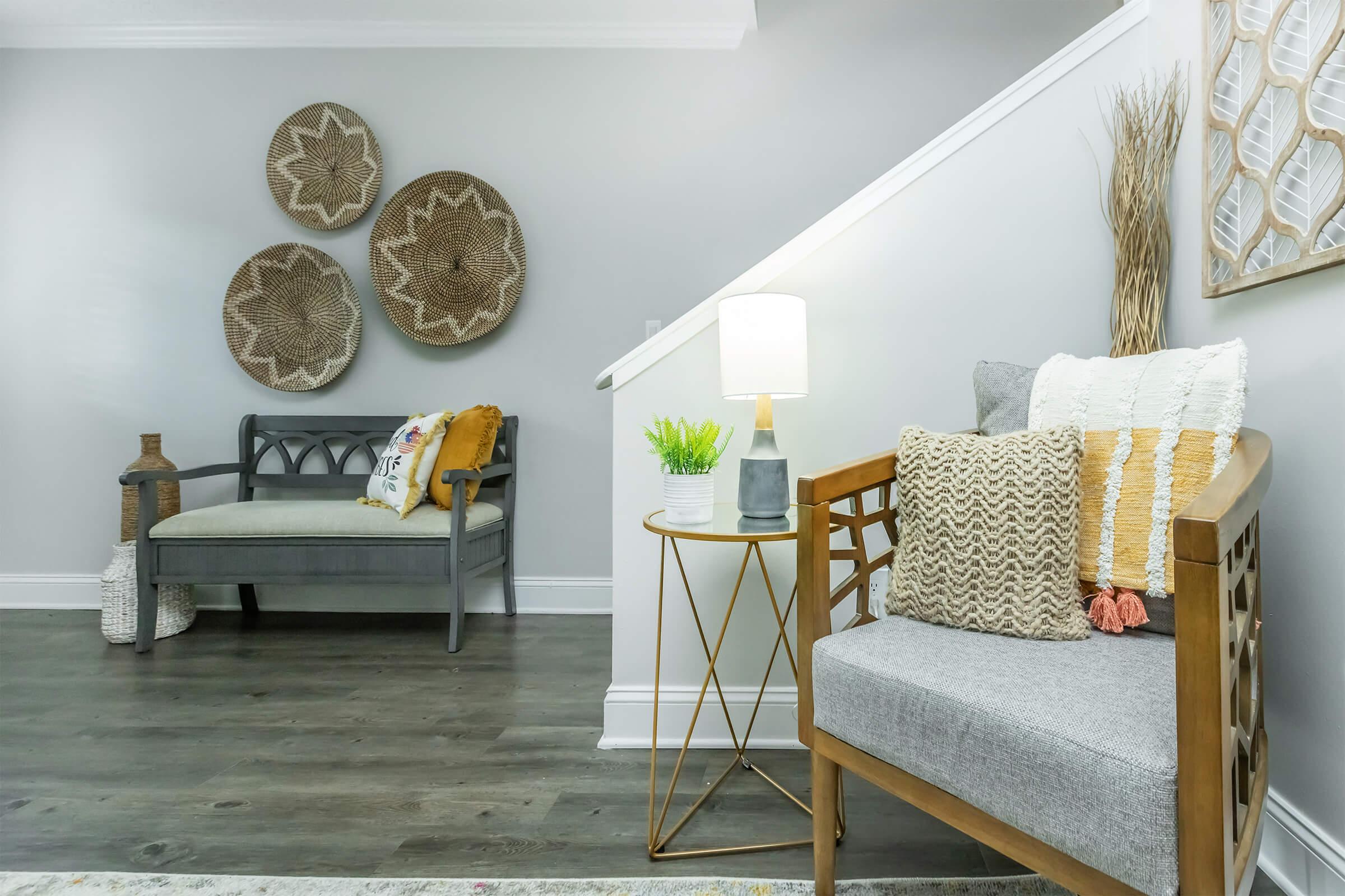A modern interior living space featuring a cozy seating arrangement. A stylish gray chair with decorative pillows is paired with a gold side table and a lamp. A plant adds a touch of greenery, and decorative woven wall art hangs above, complementing the neutral color scheme and warm wood flooring.