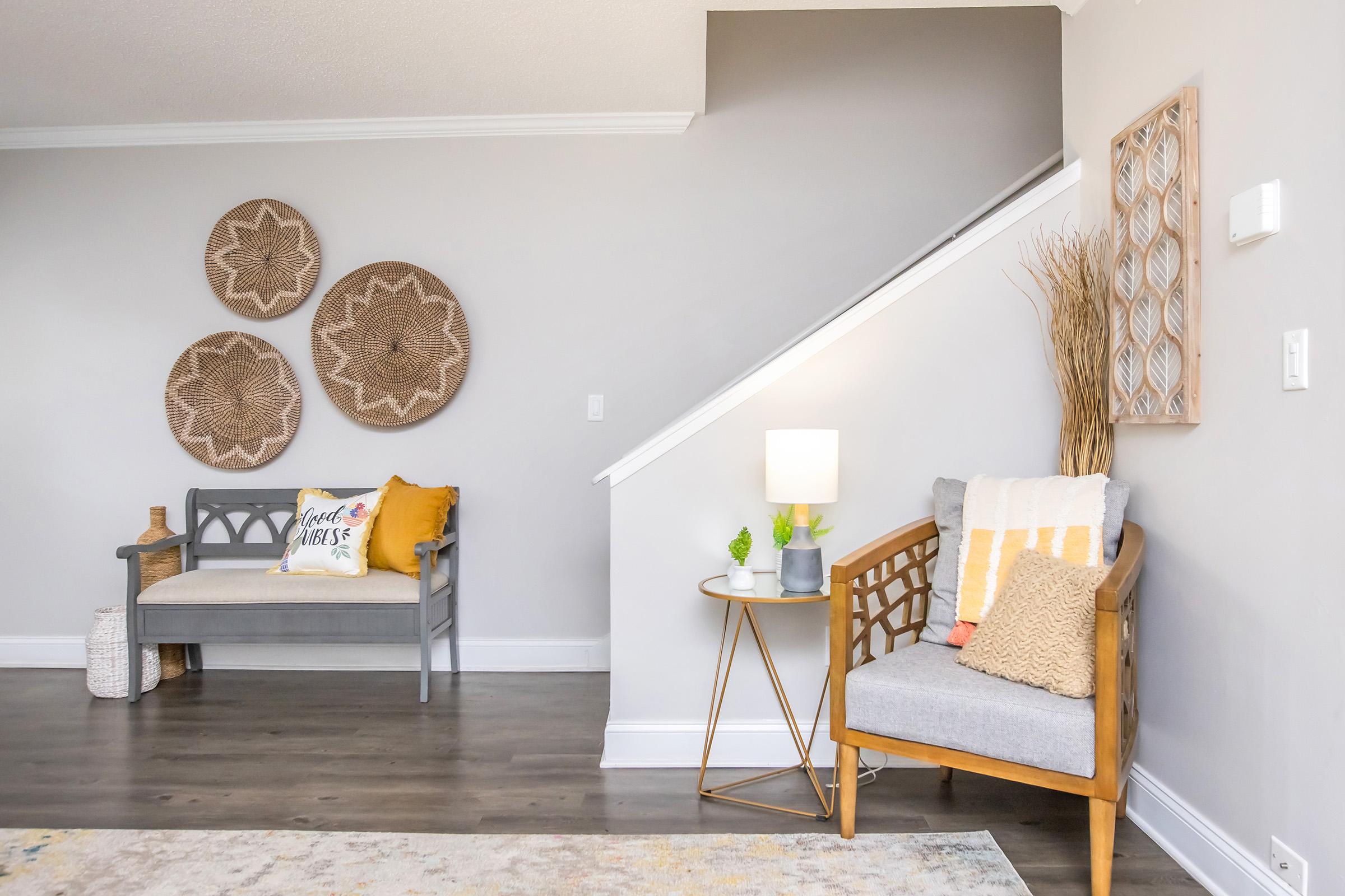 A cozy interior living space featuring a gray bench with decorative pillows, a small wooden side table with a lamp and plants, and a wooden chair with a throw blanket. The wall displays three round woven wall hangings, and there is a staircase in the background, all set in a light, neutral color palette.