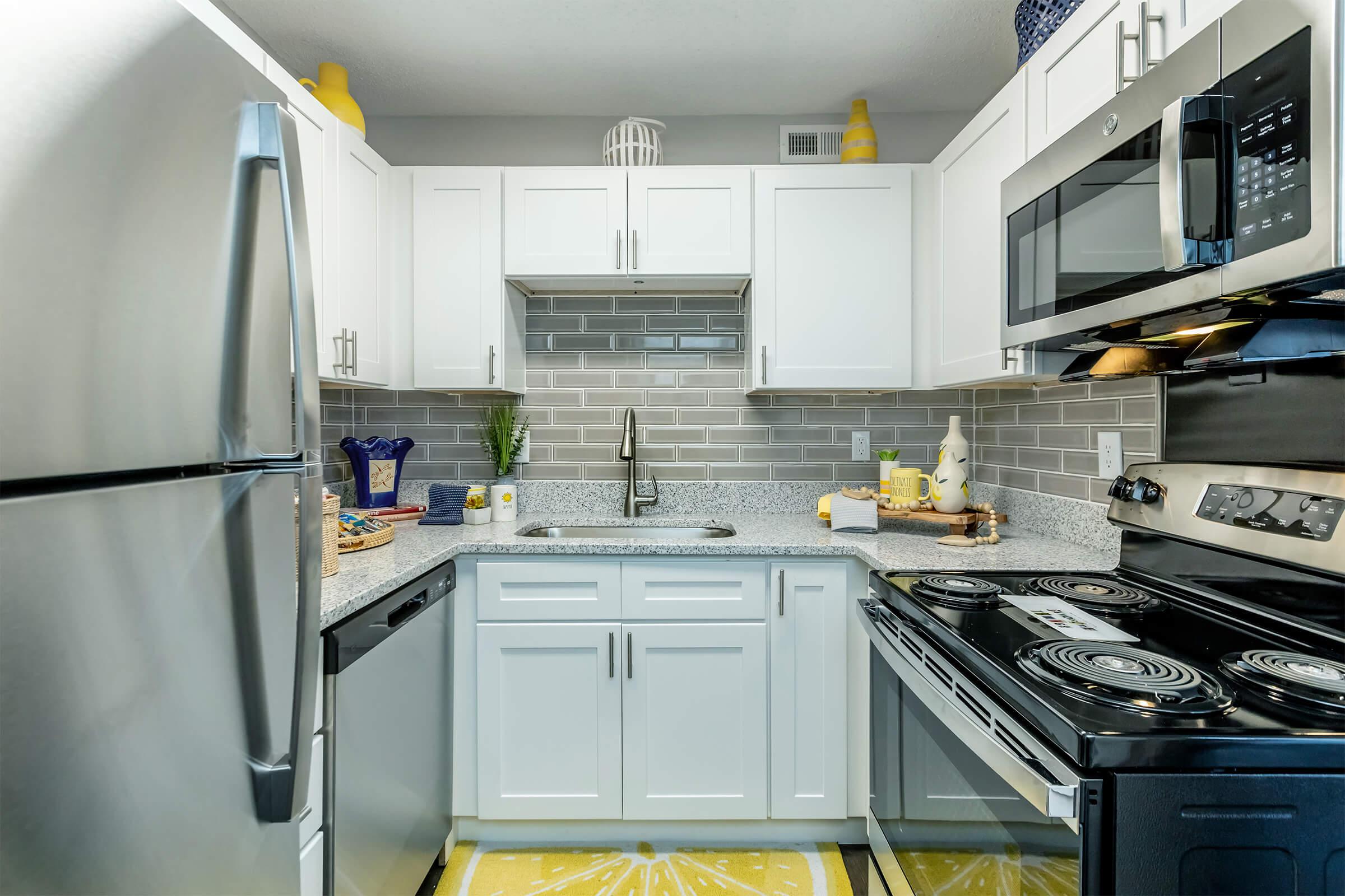 Modern kitchen featuring stainless steel appliances, including a refrigerator and oven. The cabinets are white with a gray tiled backsplash. A granite countertop with a sink is present, along with a patterned yellow rug. Items like a blue vase and kitchen utensils are neatly arranged on the countertop.