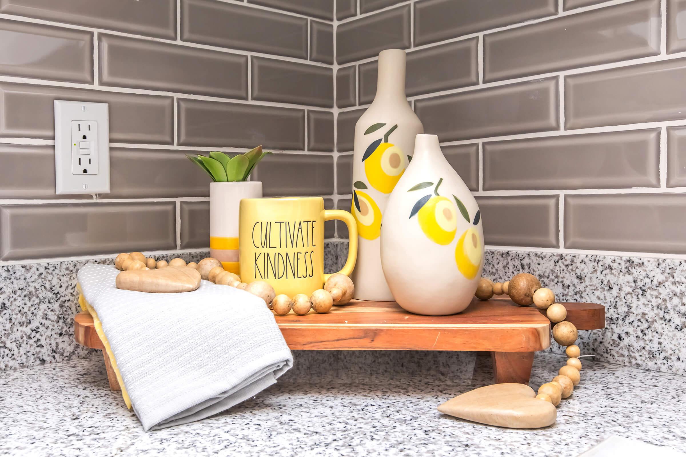 A cozy kitchen setting featuring a wooden tray with a yellow mug labeled "CULTIVATE KINDNESS," two decorative vases with a lemon design, a small green plant, a wooden heart, and a beaded garland. The background has gray subway tiles, and there's a folded dish towel nearby on the speckled countertop.