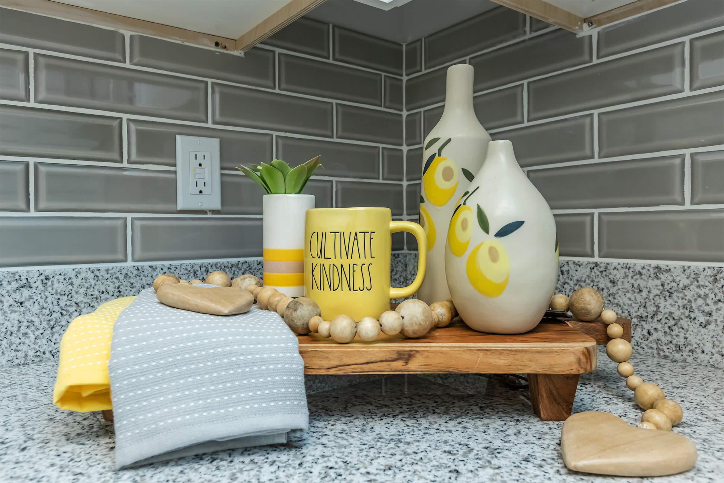 A decorative kitchen countertop featuring a wooden tray with a yellow "Cultivate Kindness" mug, two ceramic vases with lemon designs, a small green plant, a grey and yellow striped napkin, and a wooden bead garland. The background showcases a tiled backsplash with grey tiles.