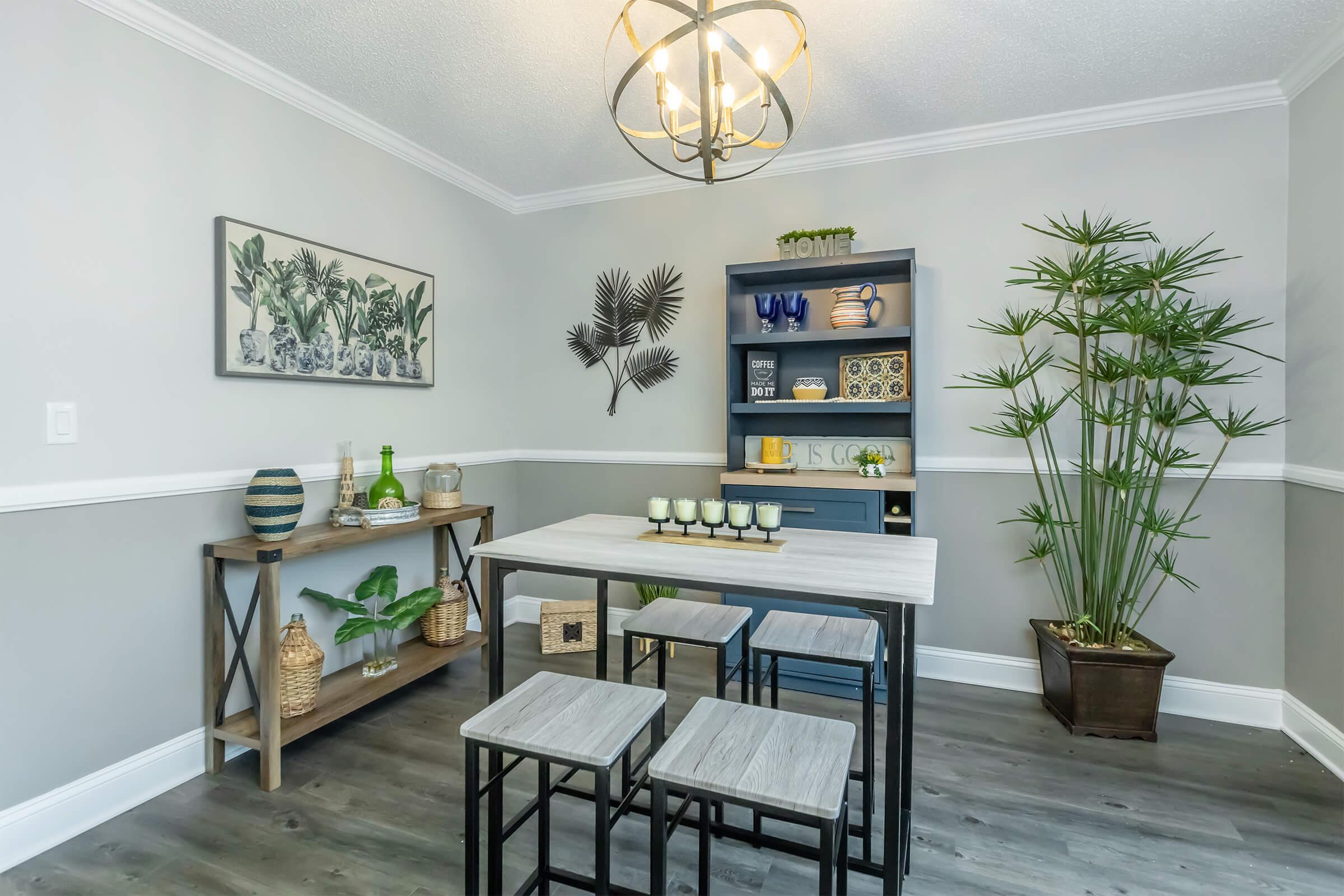 A modern dining area featuring a light wooden table with four stools, a side table displaying decorative items and plants, and a gray cabinet with colorful dishware. The walls are painted in soft gray and white, complemented by tropical-themed artwork and indoor plants, creating a fresh and inviting atmosphere.
