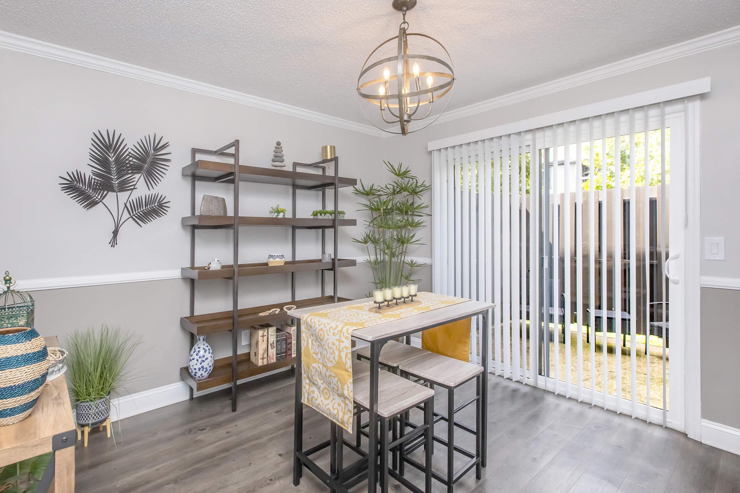 A cozy dining area featuring a wooden table with two stools, a patterned table runner, and a decorative shelf adorned with plants and decor items. A large window with vertical blinds allows natural light, and a palm leaf wall decoration adds a touch of greenery to the space.