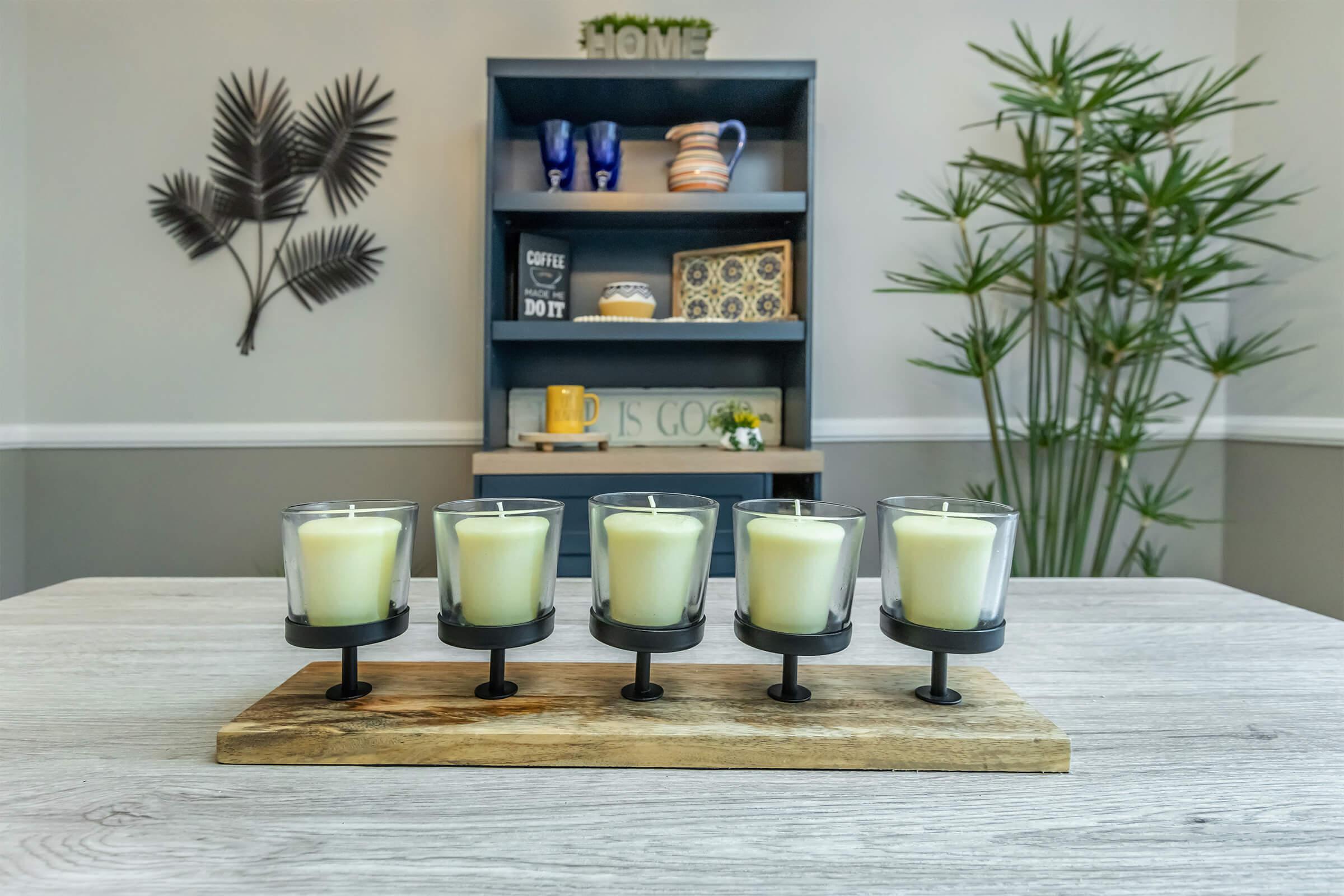 A stylish arrangement of five lit candles in glass holders on a wooden tray, displayed on a light-colored table. In the background, a modern shelf is decorated with various items, including mugs and a plant, creating a cozy home atmosphere.
