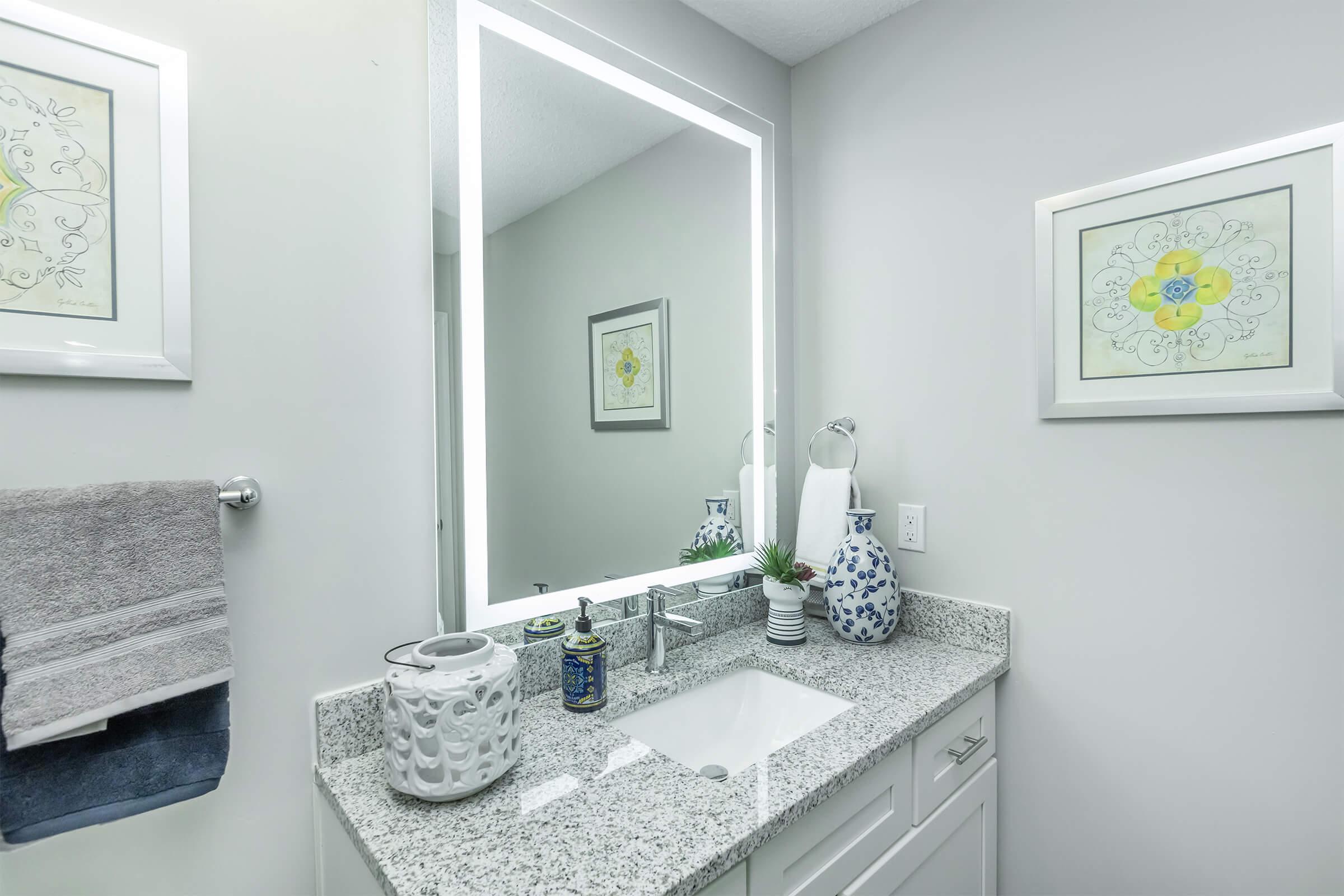 A modern bathroom featuring a granite countertop with a white sink, a backlit mirror, and decorative wall art. There are towels hanging on a rack, a blue and white vase, and various decorative items on the countertop, creating a fresh and inviting ambiance.