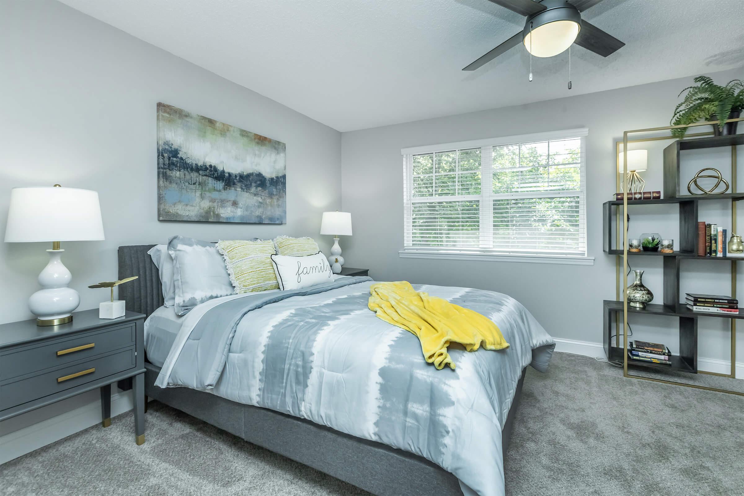 Modern bedroom featuring a gray upholstered bed with a yellow throw blanket, decorative pillows, and a light gray comforter. A stylish nightstand with a lamp sits beside the bed, and a bookshelf with plants and books adds character to the room. Natural light streams through the window, enhancing the warm ambiance.