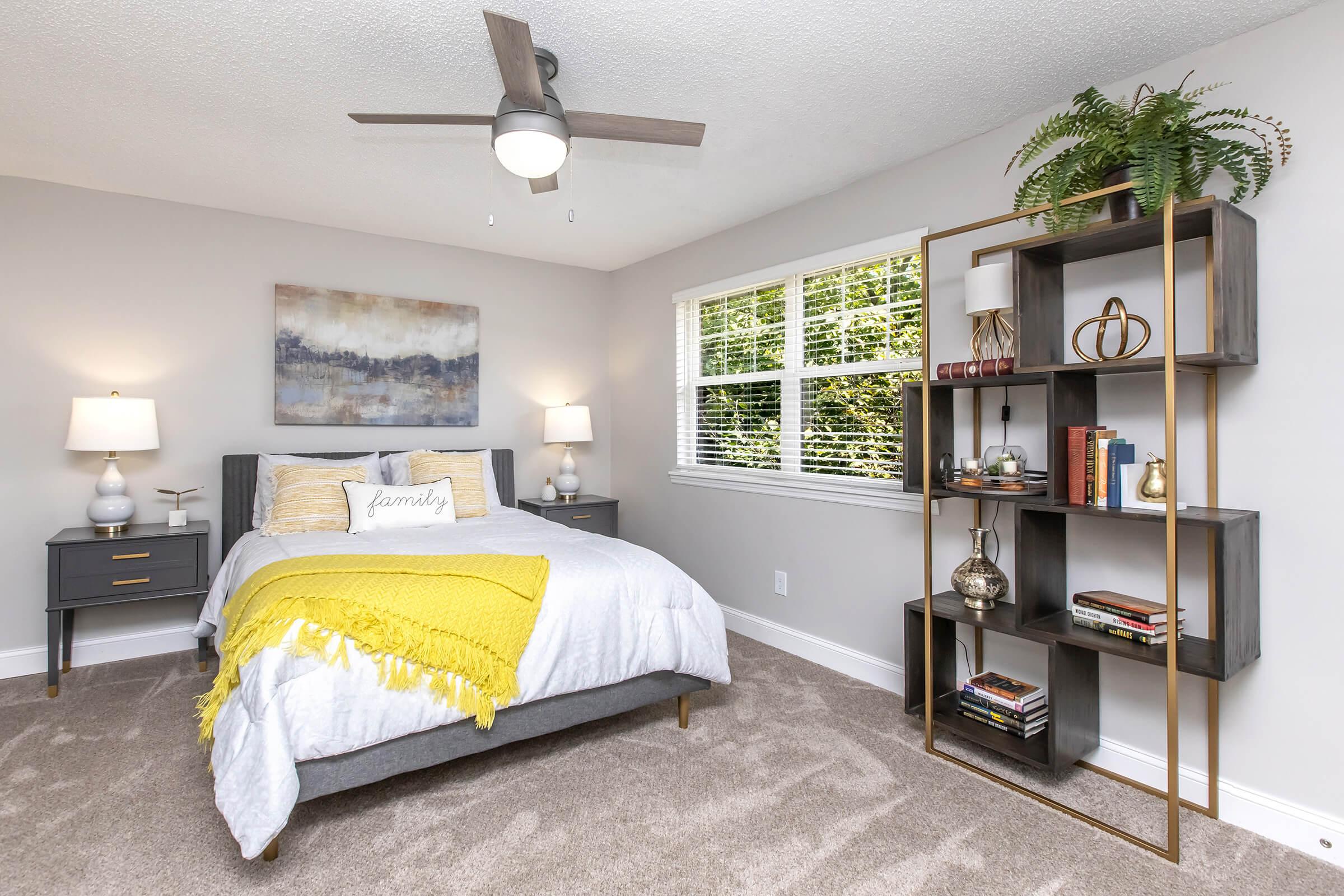 A cozy bedroom featuring a queen-sized bed with a yellow throw blanket, two bedside tables with lamps, and a large window allowing natural light. There's a modern bookshelf decorated with books and decorative items. The room has neutral walls and soft carpeting, creating a warm and inviting atmosphere.