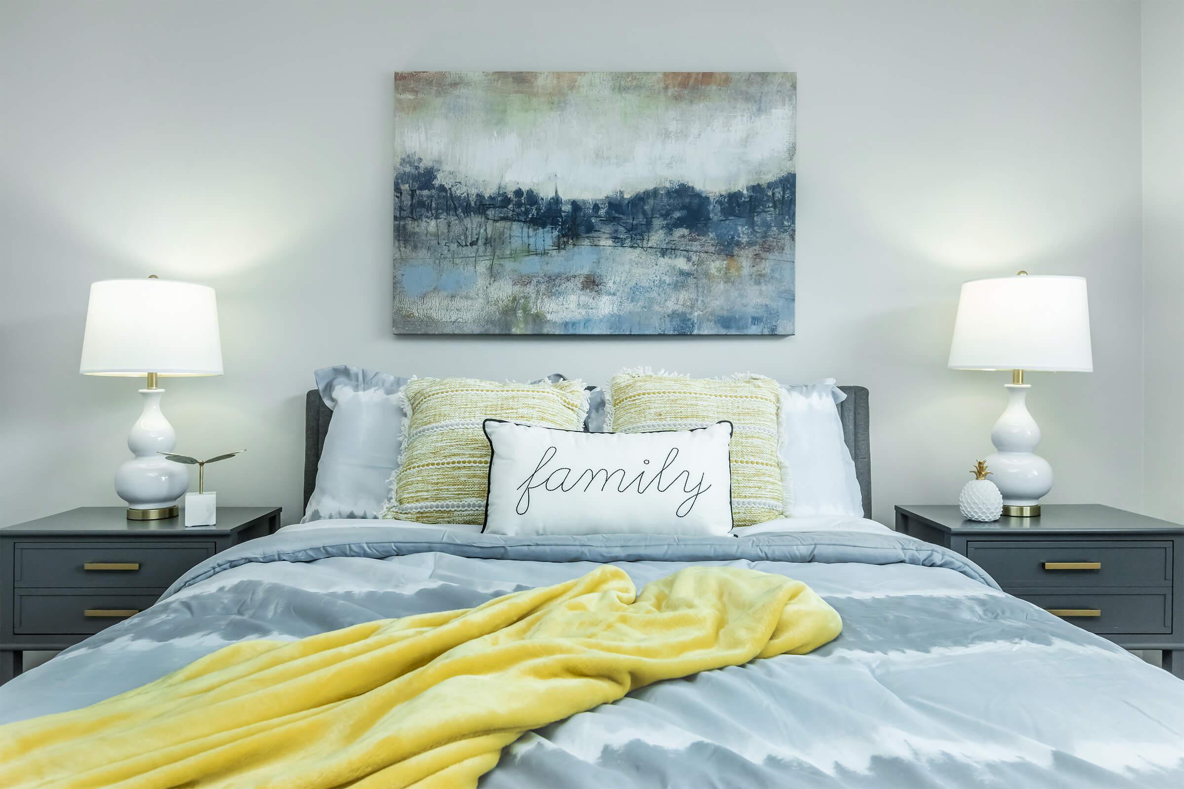 A cozy bedroom featuring a gray bed with multiple pillows, including a decorative pillow that says "family." There are two bedside tables with white lamps on each side, and a colorful abstract painting hanging above the bed. A soft yellow blanket is draped over the bed adding warmth to the space.