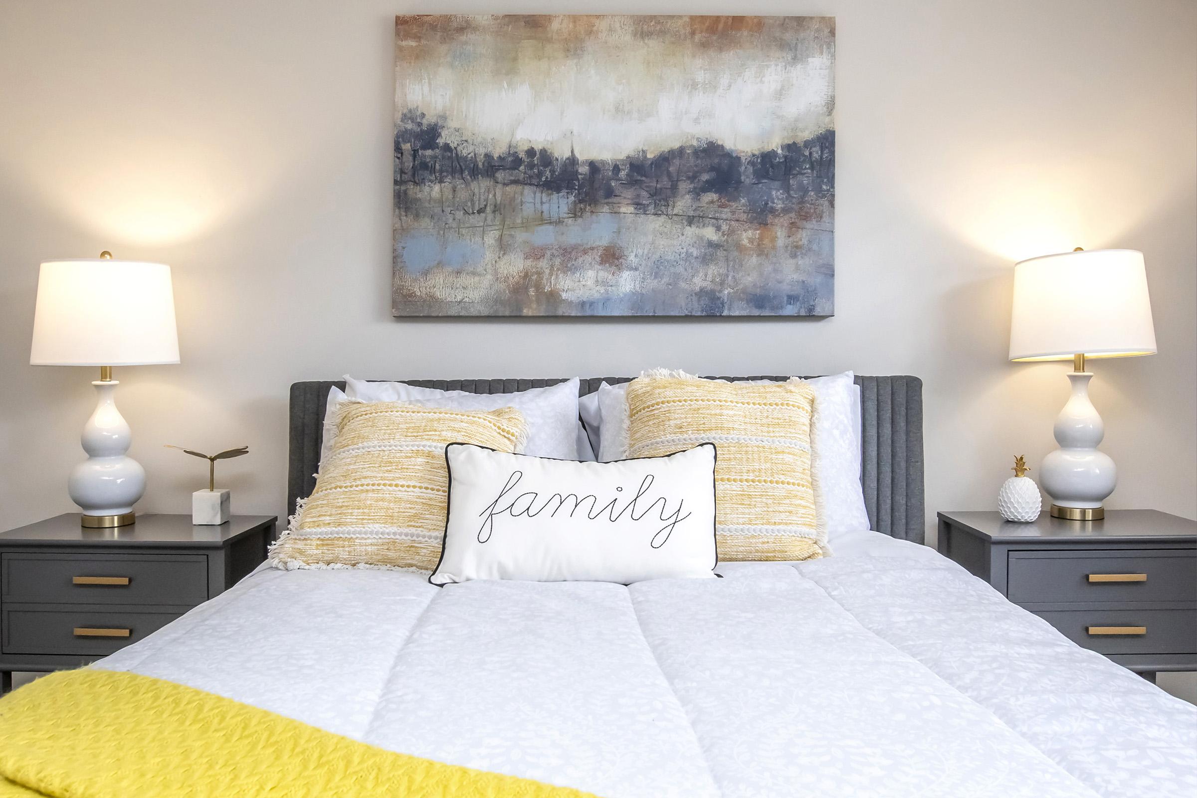 A cozy bedroom featuring a large bed with white bedding and a decorative pillow that says "family." The bed is flanked by two modern nightstands with white lamps. Above the bed hangs an abstract painting in soft colors, adding a warm touch to the space. A bright yellow throw blanket is elegantly draped over the corner of the bed.