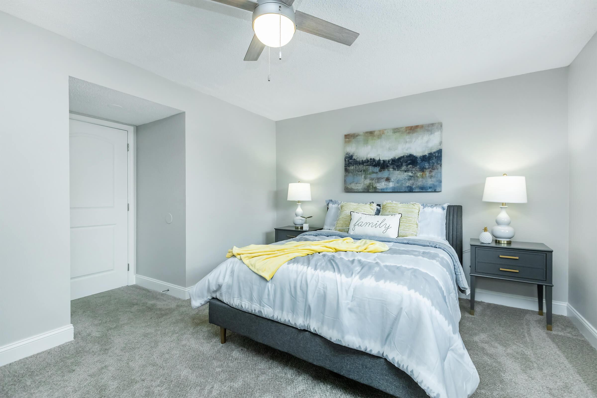 A cozy bedroom featuring a gray upholstered bed with a light blue and white comforter, yellow throw blanket, and decorative pillows. Two bedside tables with lamps are positioned on either side of the bed, and a piece of abstract wall art hangs above. The walls are painted in a soft gray, and the room has a carpeted floor.