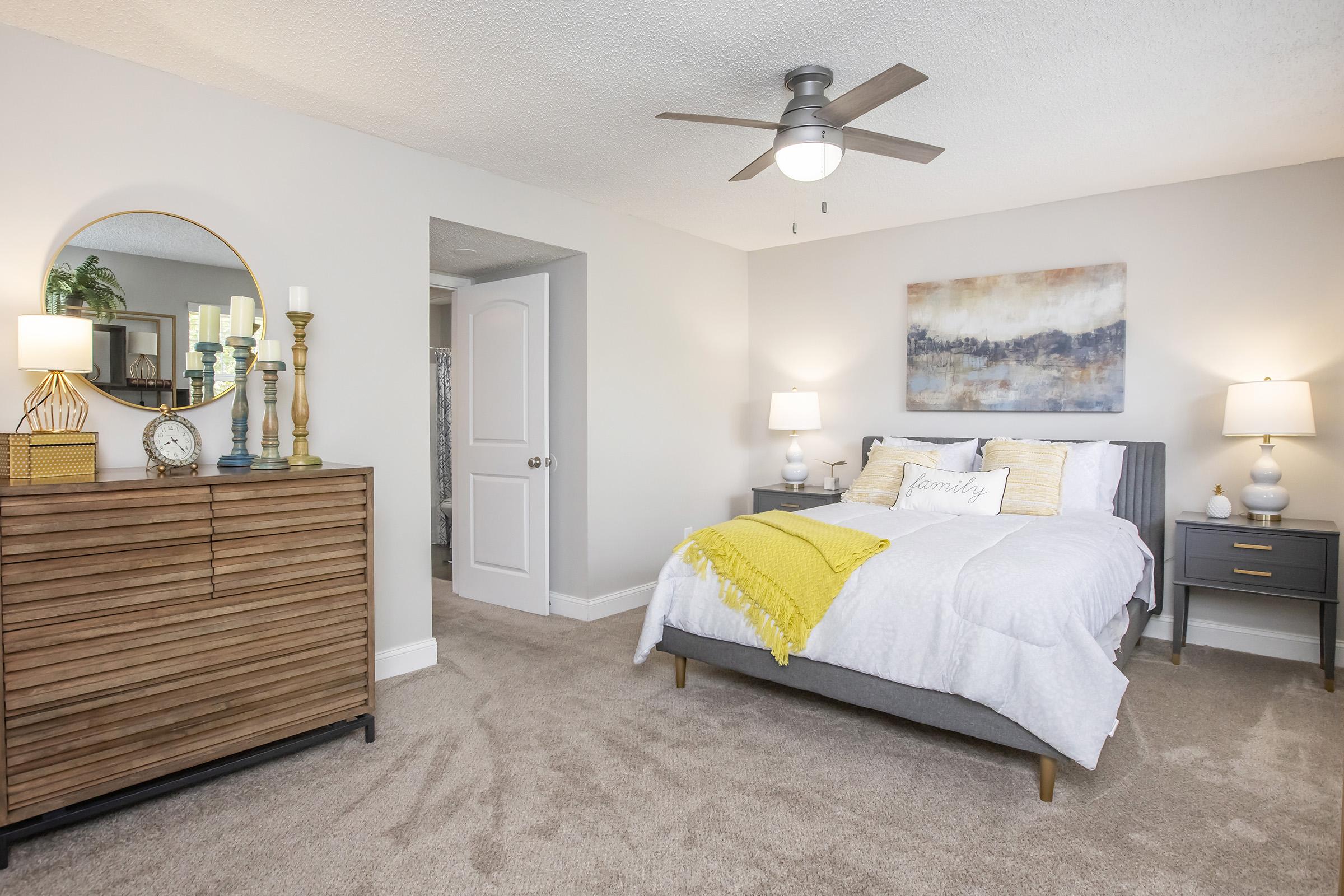 A cozy bedroom featuring a grey bed with white bedding and a yellow throw blanket. There is a wooden dresser with decorative items and a round mirror above it. On either side of the bed are nightstands with lamps. A colorful abstract painting hangs on the wall, and a doorway leads to another room. The flooring is carpeted.