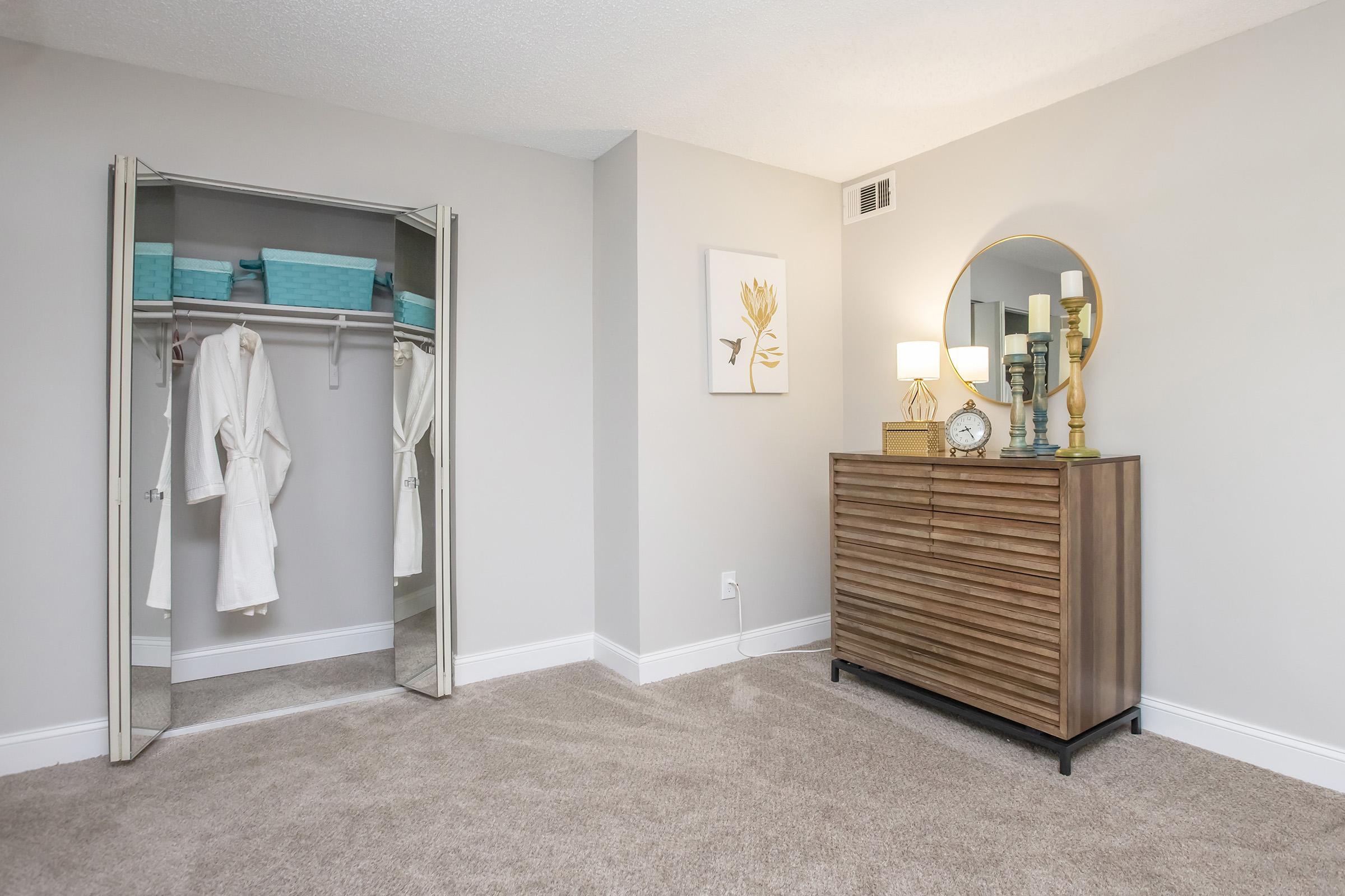 A cozy bedroom featuring a mirrored closet with white robes hanging inside, a wooden dresser with decorative items and a round mirror above it. Soft, neutral walls and carpet create a serene atmosphere. A piece of art with a floral design is hung on the wall, adding a touch of elegance to the space.