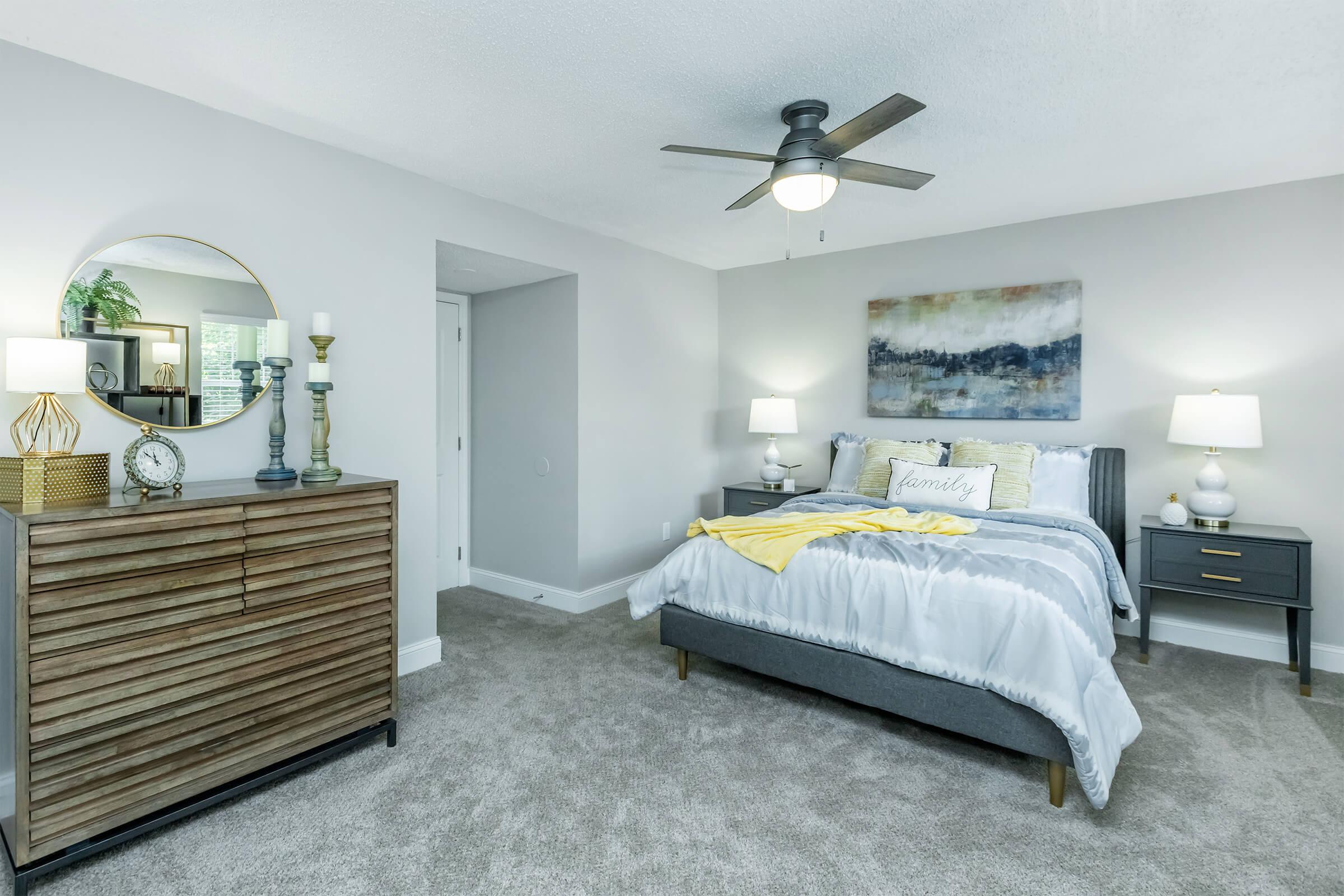 A stylish modern bedroom featuring a gray upholstered bed with a white and yellow bedding set, two bedside tables with lamps, a wooden dresser, and a round mirror. The walls are painted light gray, and there is a painting above the bed. The carpet is plush, creating a cozy atmosphere.