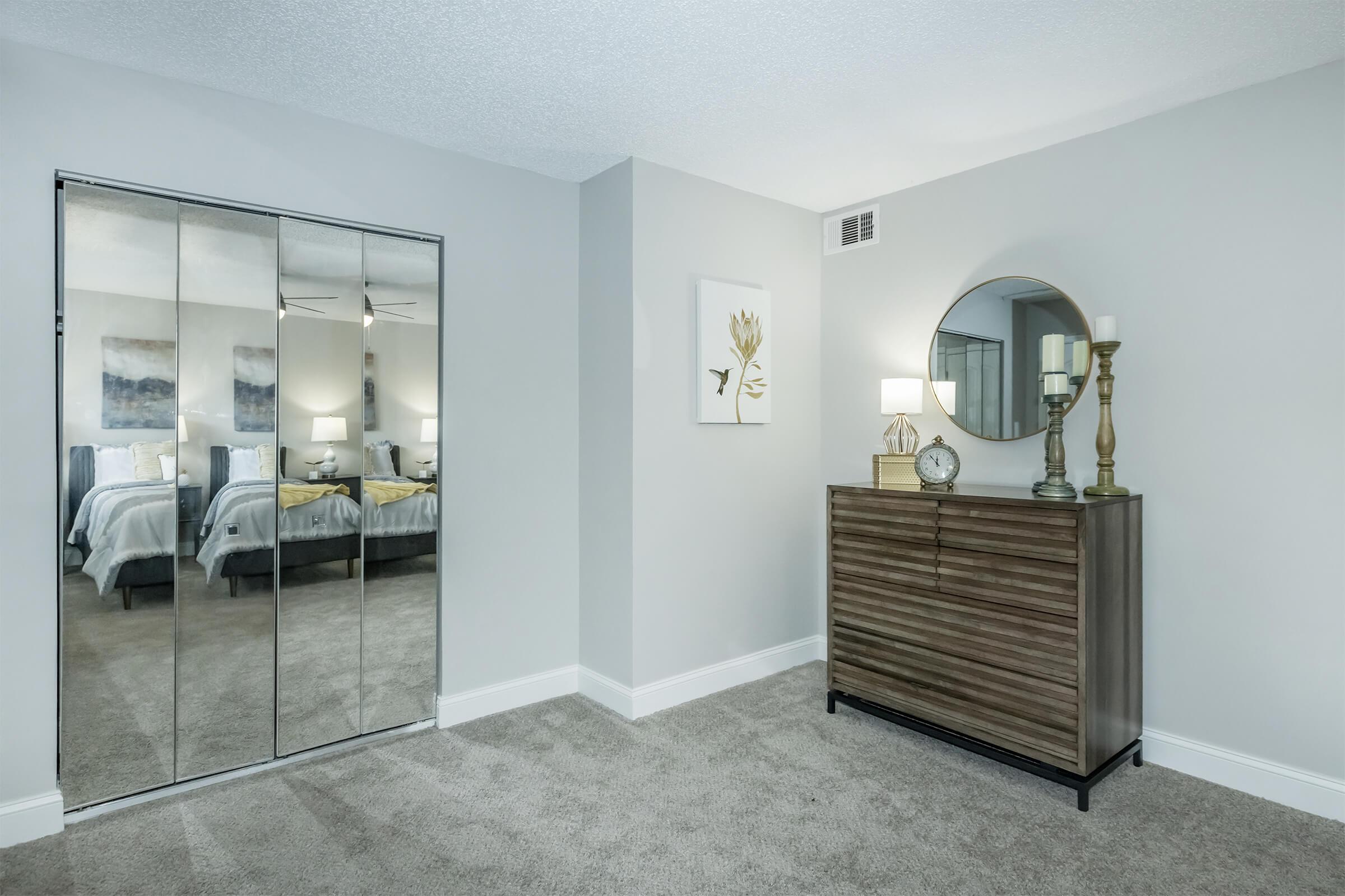 A stylish bedroom corner featuring a modern dresser with decorative items, a round mirror, and artwork on the wall. A sliding mirror closet reflects the neatly made bed and soft lighting, creating a cozy and inviting atmosphere. The color palette includes neutral tones and soft textures.