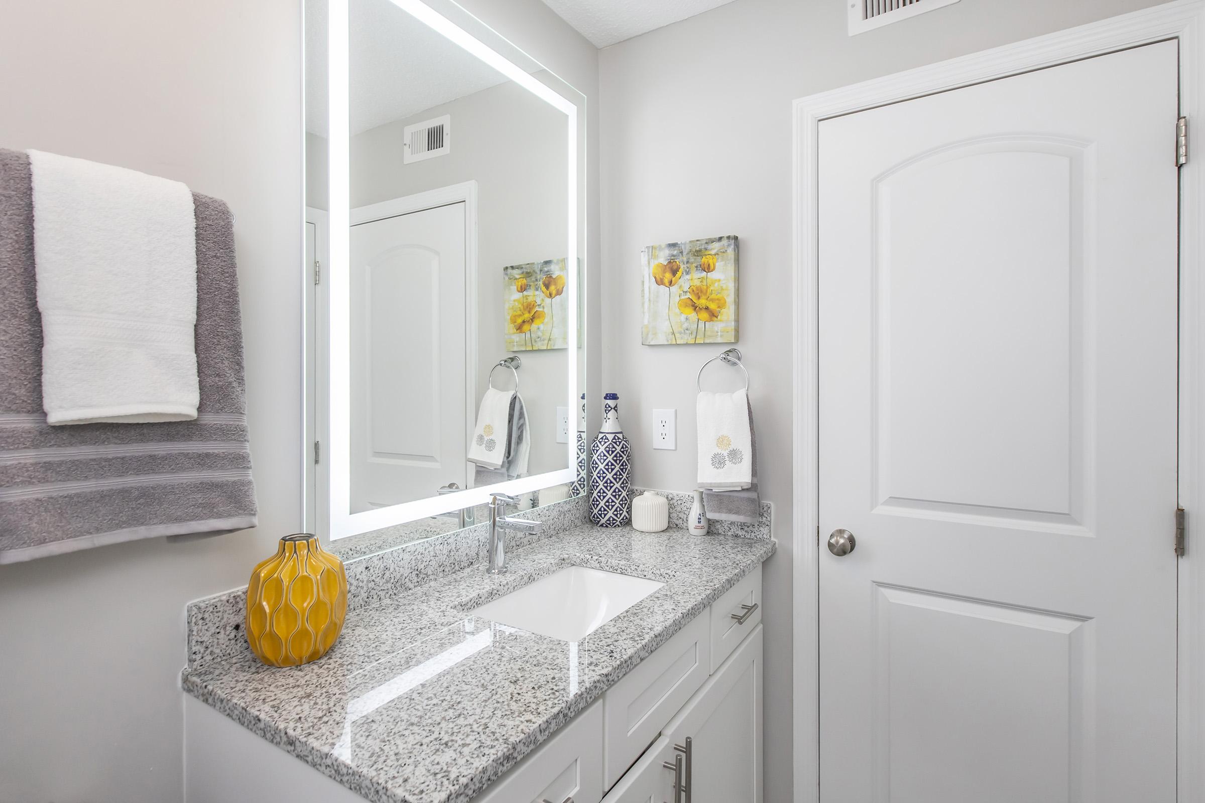 A modern bathroom featuring a gray and white color scheme, a granite countertop with a white sink, and yellow decorative elements. The walls are adorned with floral artwork, and a backlit mirror reflects the space. A towel rack holds a gray towel, while a door leads to another area.