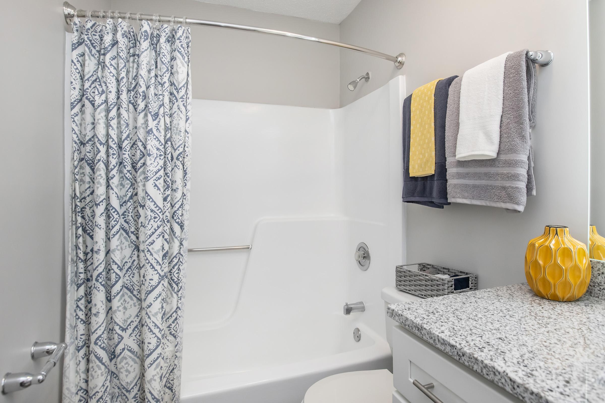 A modern bathroom featuring a white bathtub with a shower curtain displaying a blue and white pattern. There are neatly arranged towels in gray, white, and yellow hanging on a chrome rack. A stylish yellow vase sits on a granite countertop, alongside a small gray basket, creating a clean and inviting space.