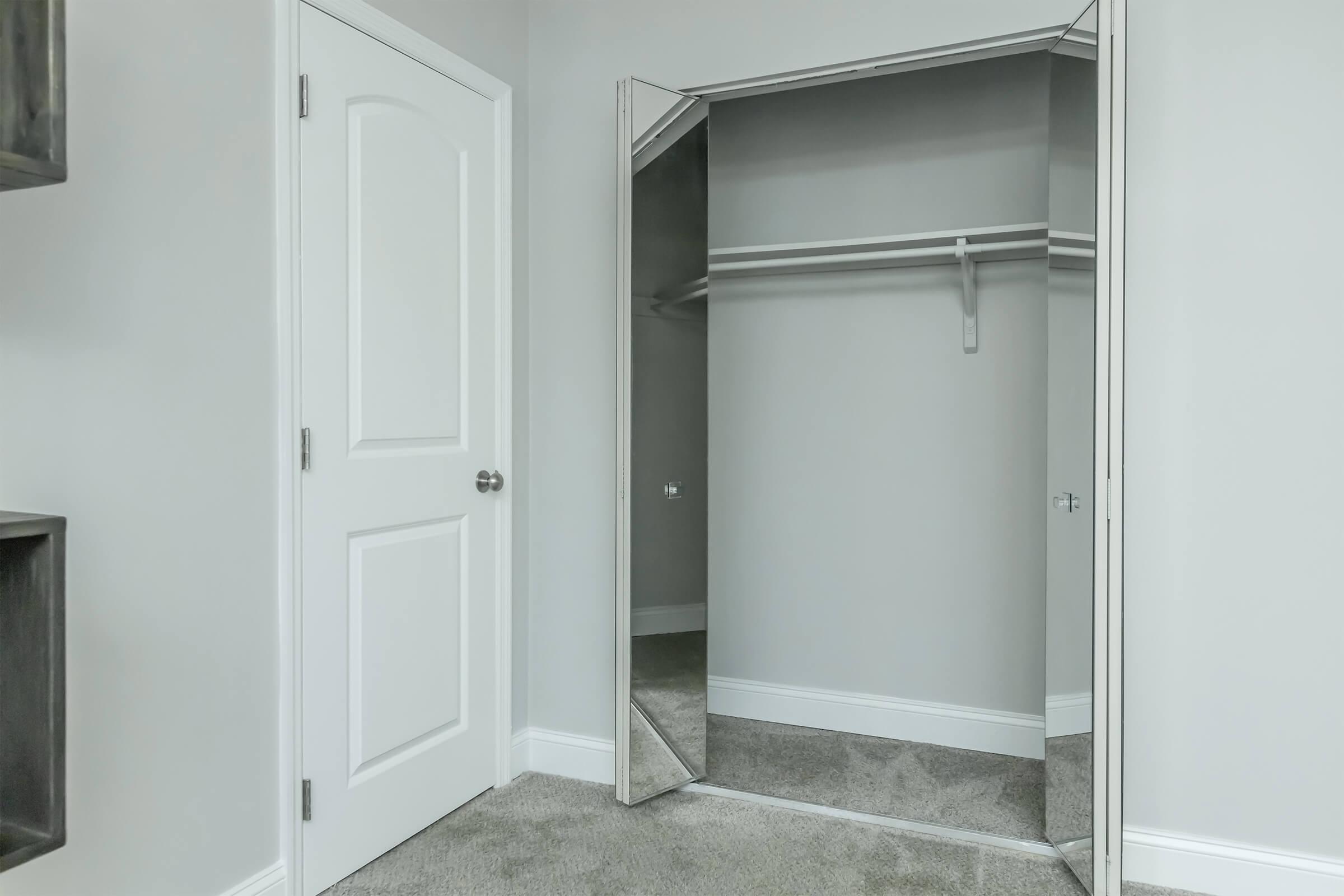 A modern closet with mirrored doors, revealing an empty space inside. There is a single clothes rod for hanging items. The walls are painted light gray, and the floor is carpeted in a neutral shade. A white door is visible to the left.