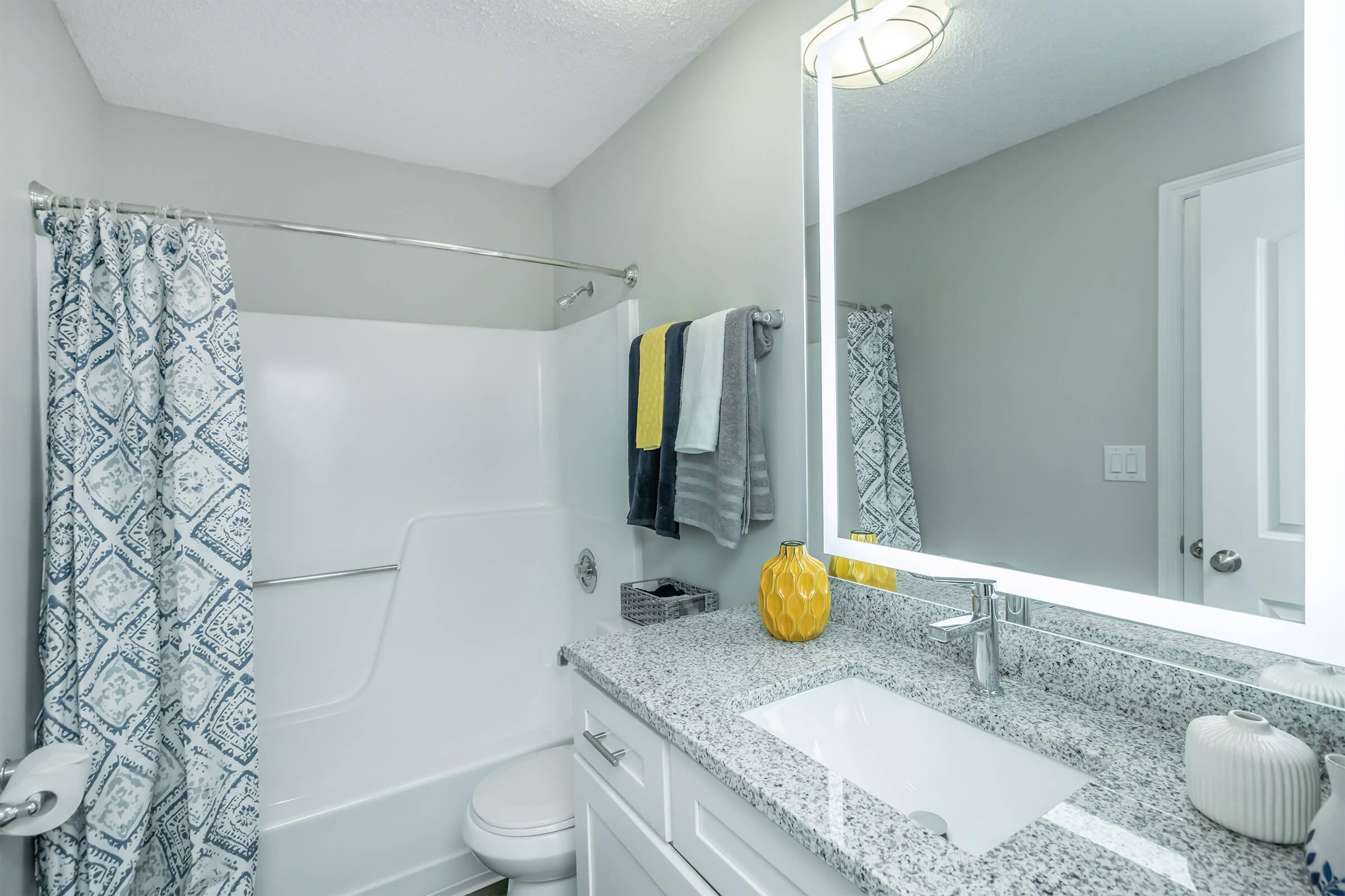 Bathroom featuring a modern design with gray walls, a white bathtub with shower curtain, and a granite countertop. A large mirror is above the sink, which has a contemporary faucet. There are neatly arranged towels in various colors on a towel rack, and a decorative yellow vase on the countertop adds a pop of color.