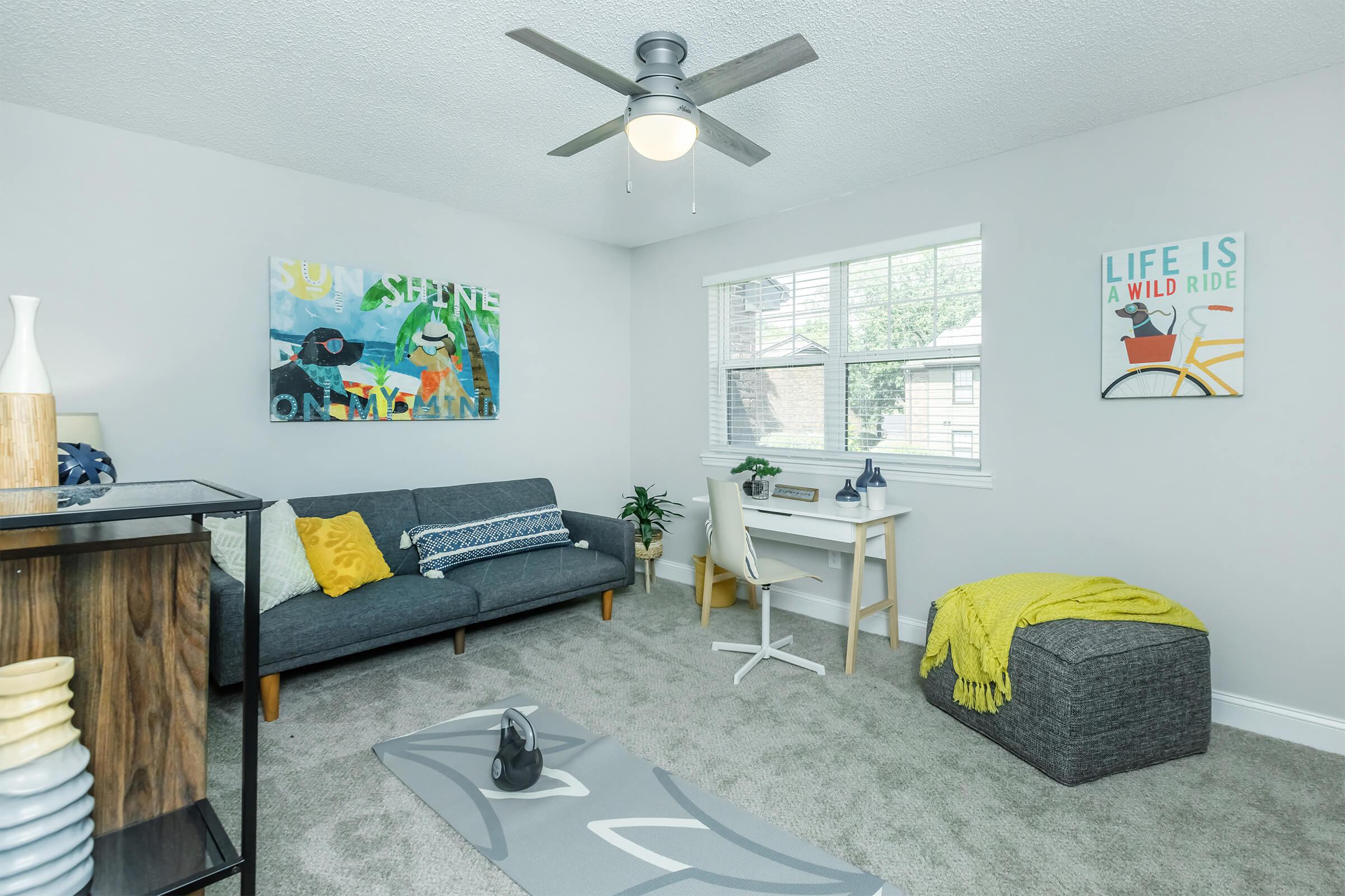 A modern, well-lit living room featuring a gray sofa with colorful throw pillows, a small dining table and chairs, artwork on the walls, a plant near the window, a cozy yellow throw blanket, and a yoga mat on the floor. The room has a ceiling fan and light-colored walls, creating a fresh atmosphere.
