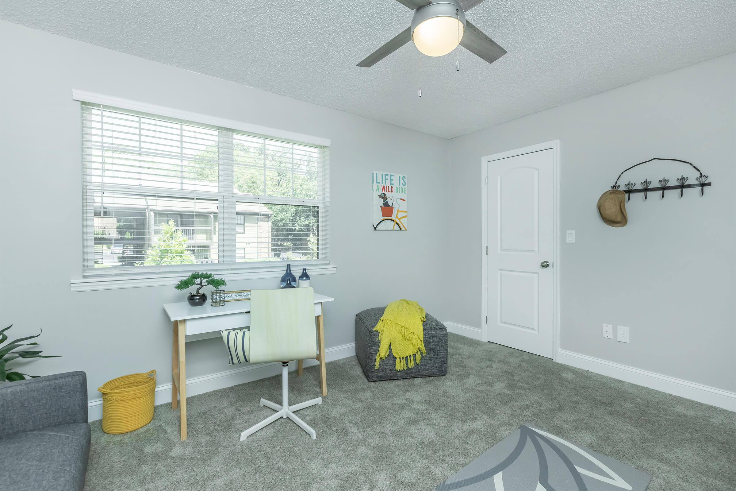 A bright and modern room featuring a small desk with two decorative items, a cozy bean bag chair, and a soft rug. Natural light fills the space through a window adorned with blinds. The walls are painted light gray, and there's a cheerful wall art piece that reads "LIFE IS." A yellow throw blanket adds a pop of color.