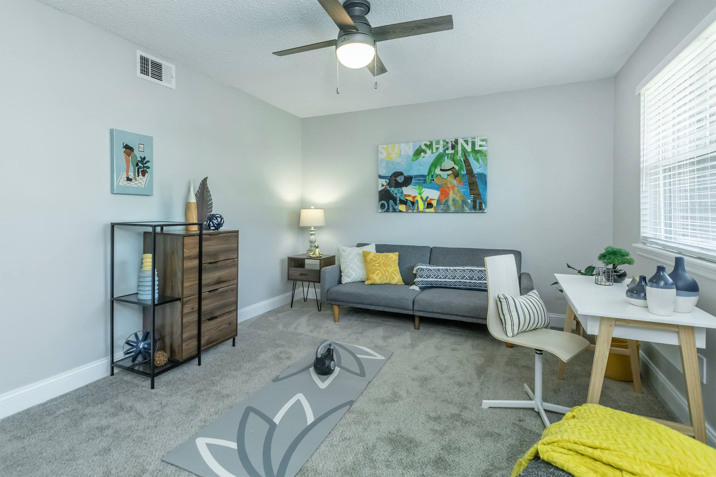 A cozy living room featuring a gray sofa with colorful cushions, a wooden dresser, a small dining table with chairs, and decorative art on the walls. The room has a ceiling fan and a neutral color palette, creating a bright and inviting atmosphere with natural light from the windows.