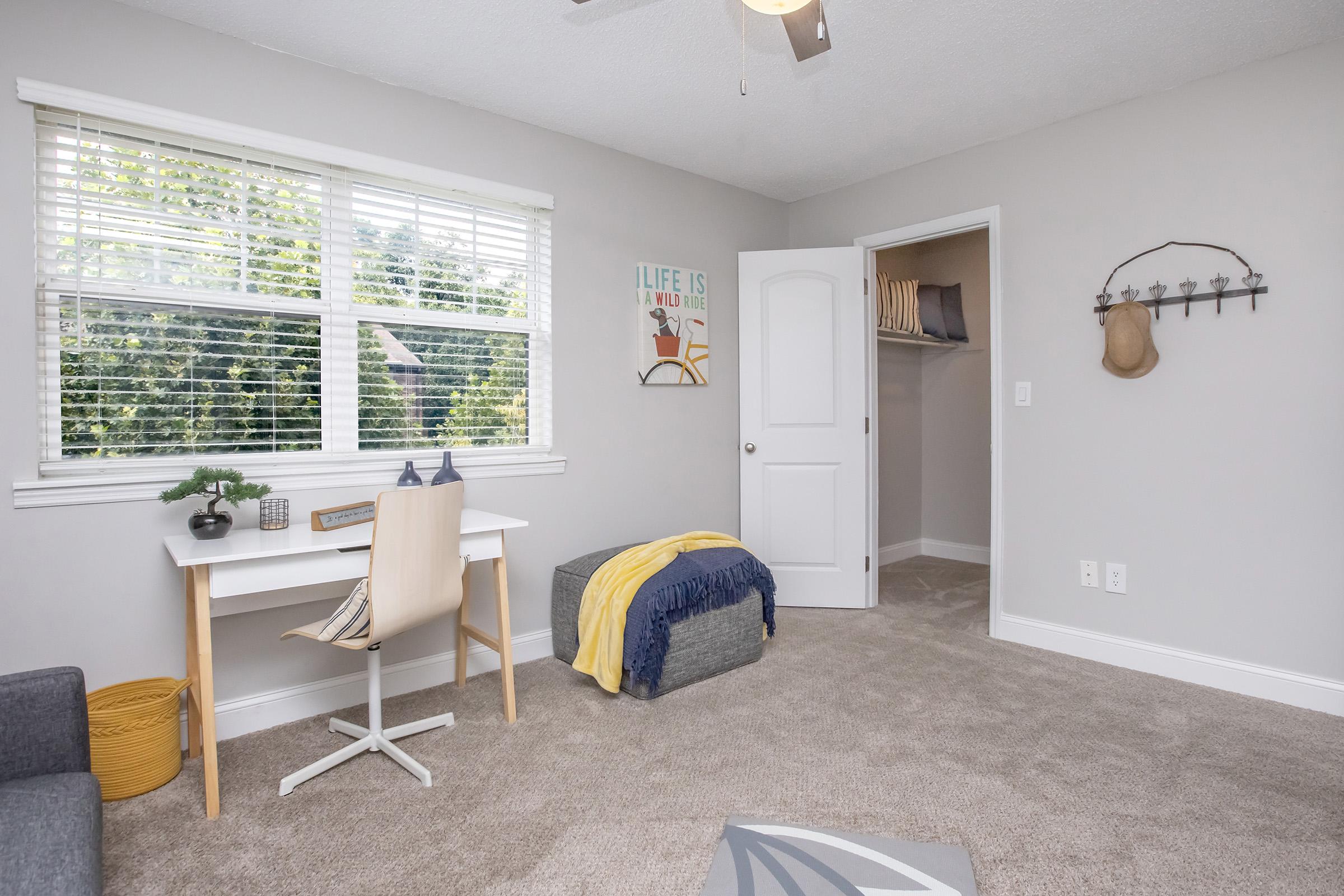 A modern, brightly lit bedroom featuring a desk with a chair, a gray ottoman, and a beige blanket. There’s a window with blinds, a closet door, and wall decor with the text "LIFE IS." The room has neutral-colored walls and carpet, creating a cozy atmosphere.